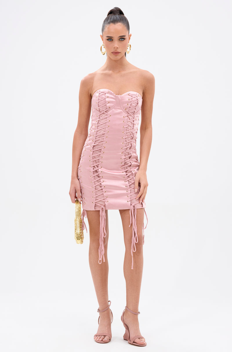 A woman wears the FAR FROM CASUAL SATIN MINI DRESS, a strapless blush pink mini with corset-style lace-up front and sides, paired with a gold beaded clutch, pink heeled sandals, and hoop earrings against a white background. 4
