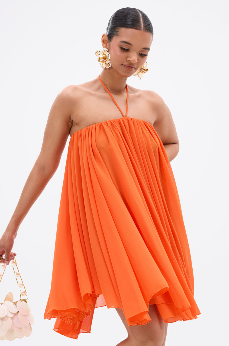 A woman in the ARIANA CHIFFON SHIFT MINI DRESS, a strapless orange halter-neck style, stands against a white background with a light bag and large gold statement earrings. 1