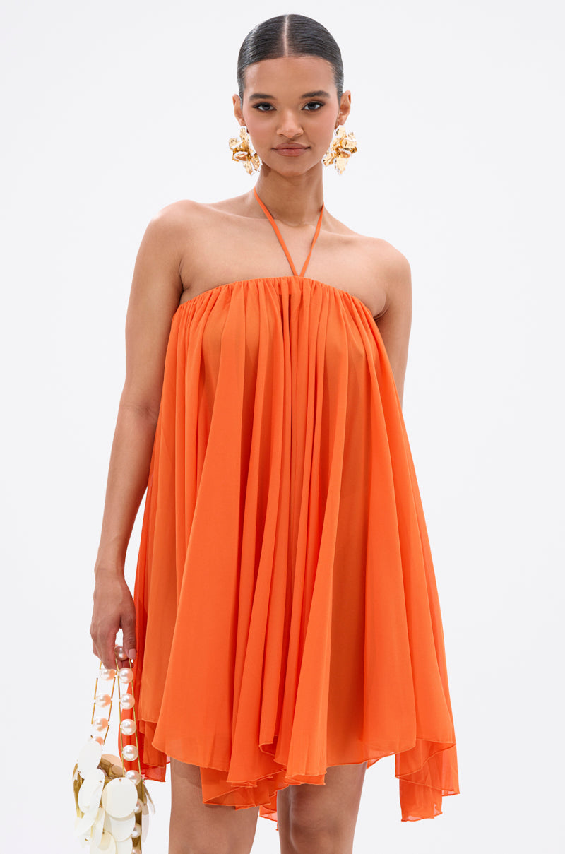 A woman wears the ARIANA CHIFFON SHIFT MINI DRESS with a halter neck strap, paired with a white and gold handbag and bold gold earrings. Her slicked-back hair and confident expression complete her chic ensemble. 2