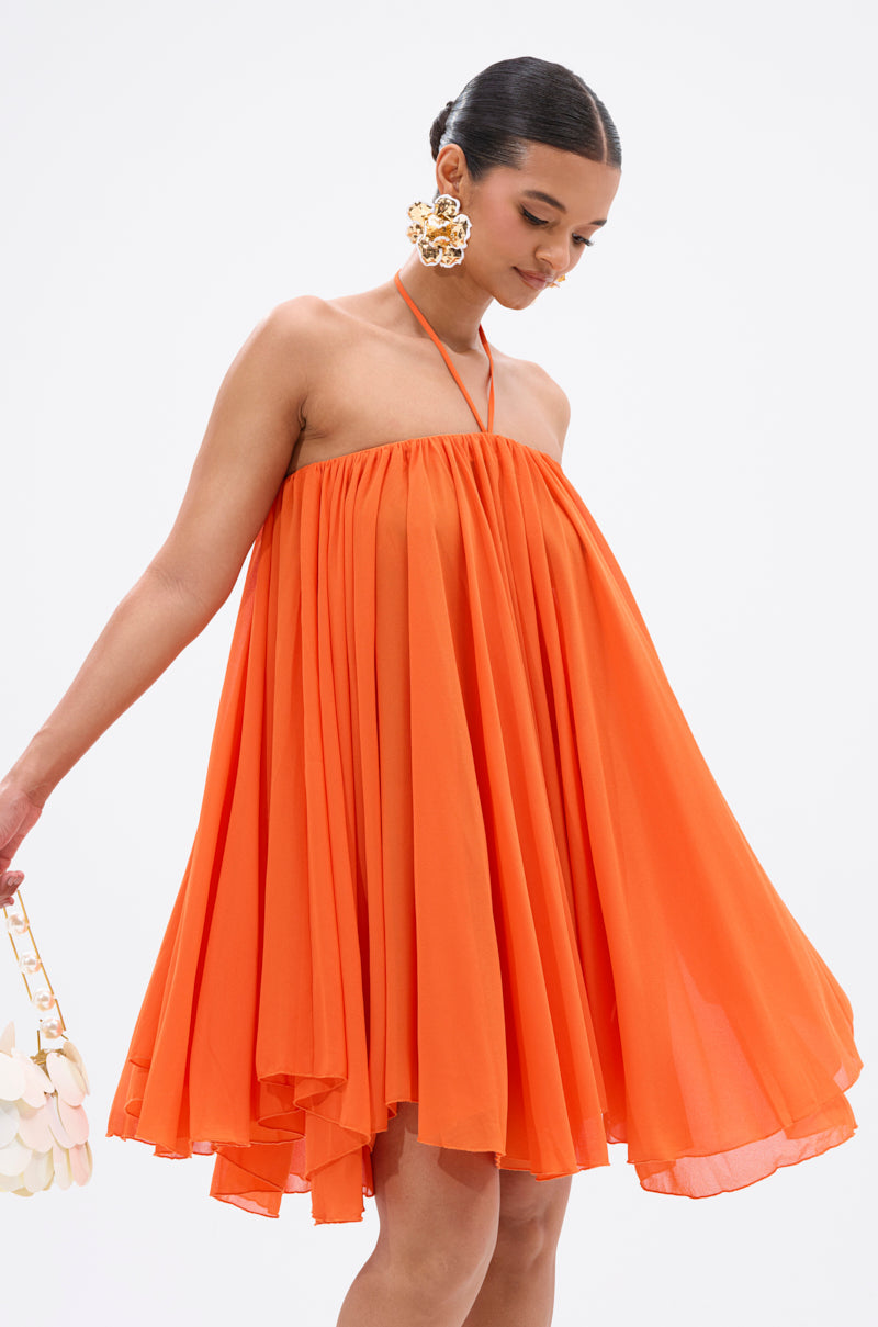 A woman wears the bright orange ARIANA CHIFFON SHIFT MINI DRESS with a halter neckline, slicked-back hair, large floral earrings, and holds a white handbag with circular accents against a plain white background. 3