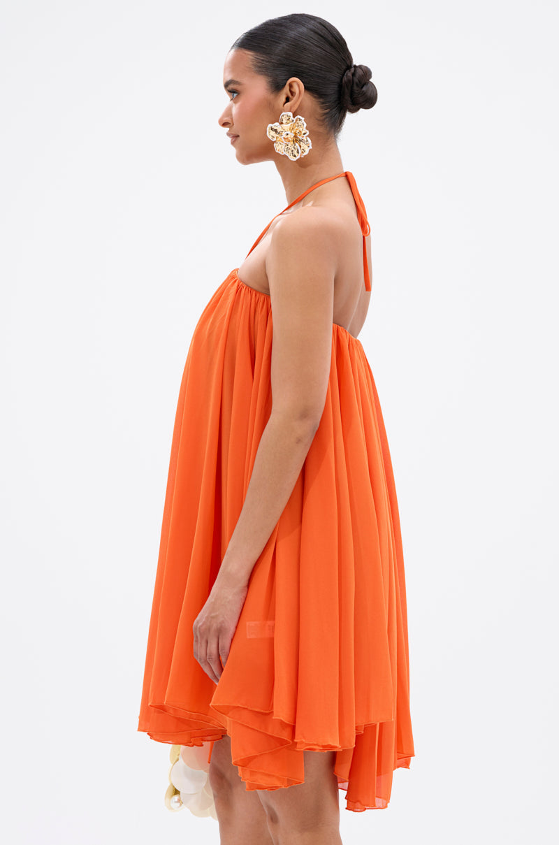 A woman in profile wears the flowy, bright orange ARIANA CHIFFON SHIFT MINI DRESS with a halter neck, large floral earrings, a neat low bun, and holds a white clutch against a plain white background. 4