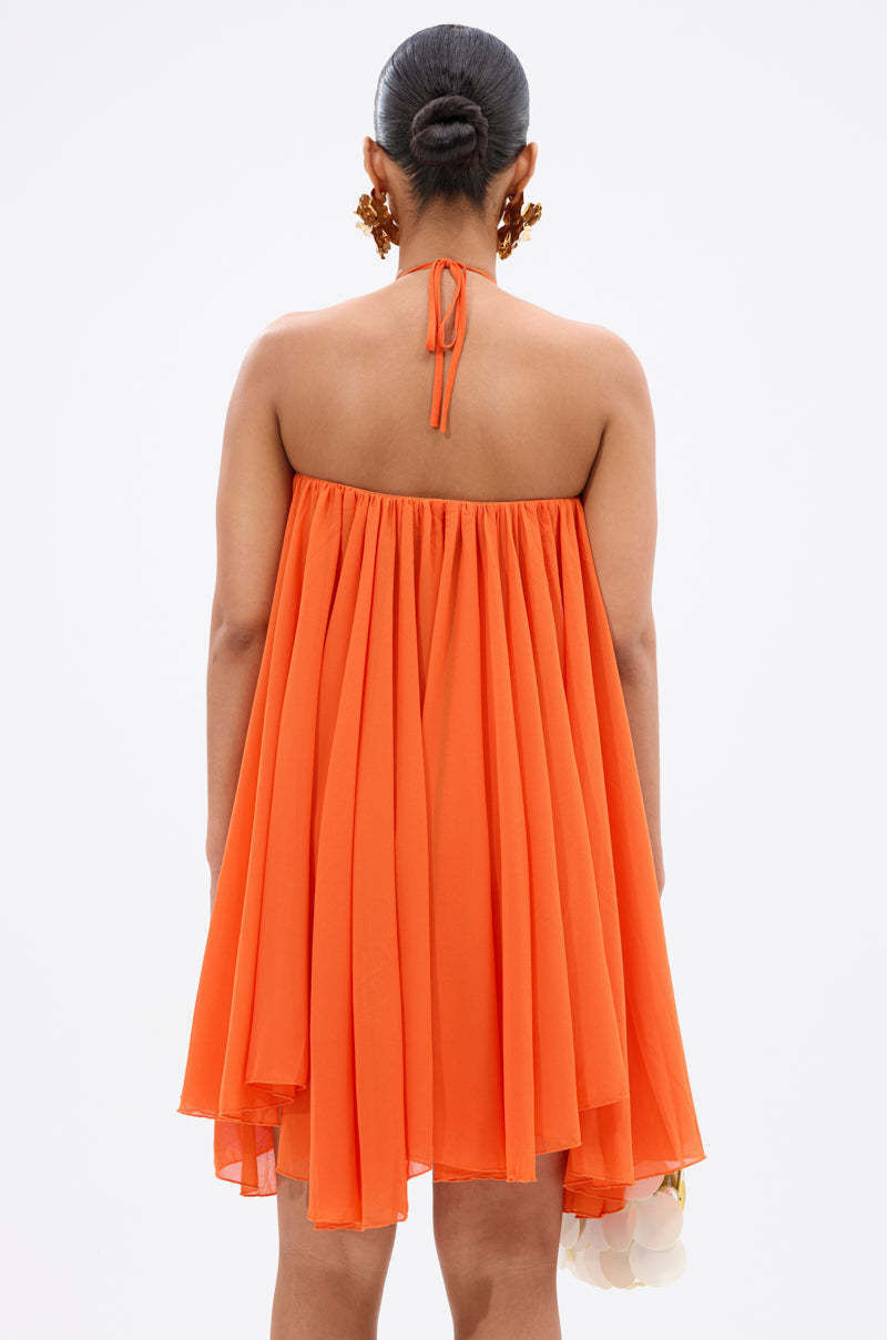 A woman faces away, wearing the vibrant orange ARIANA CHIFFON SHIFT MINI DRESS with a flowy halter neck and tie detail. Her hair is in a bun, and she holds a white item at her side against a plain white background. 5