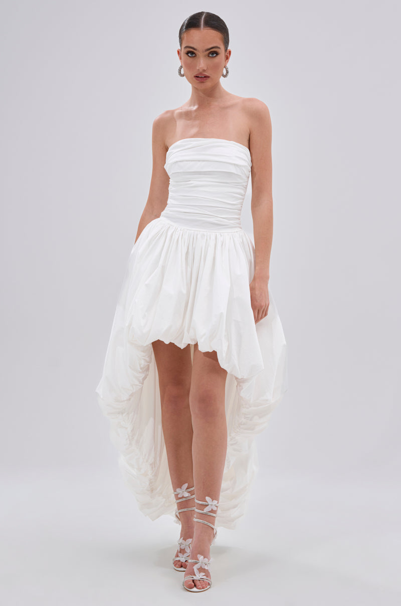A woman stands against a plain background in the THIS AND THAT POPLIN HIGH LOW DRESS, a strapless cotton poplin design with a ruched bodice and high-low hem, styled with white floral-embellished heels and statement earrings.