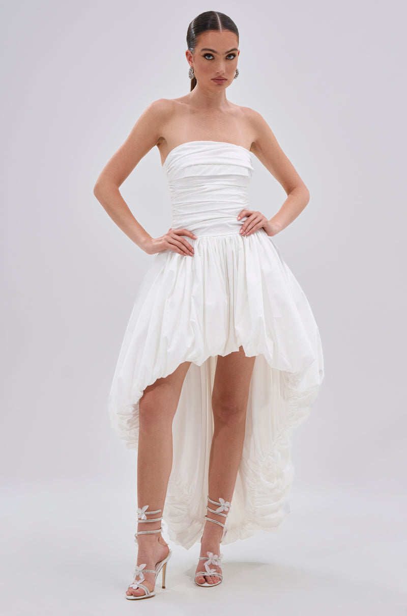 A woman poses in the THIS AND THAT POPLIN HIGH LOW DRESS, featuring a strapless fitted bodice and voluminous hem, styled with white floral heeled sandals, set against a plain light background.