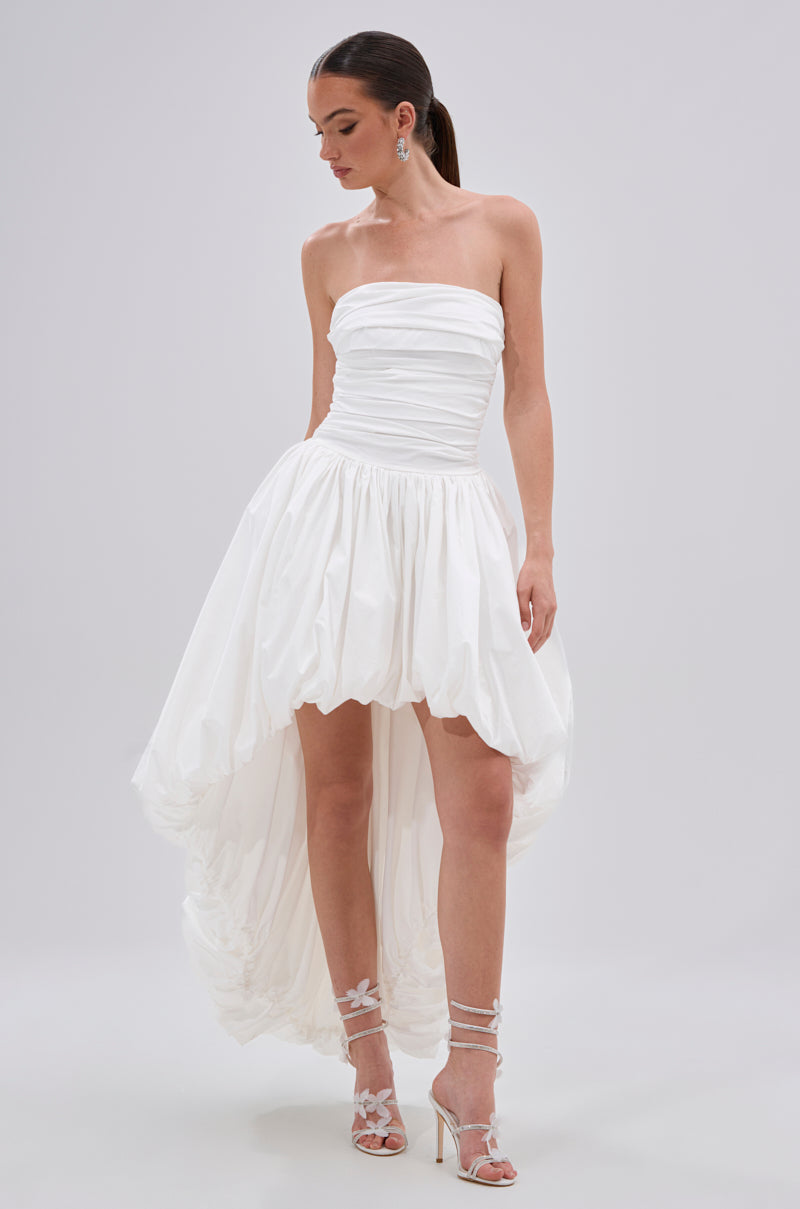 A woman wears the THIS AND THAT POPLIN HIGH LOW DRESS, featuring a strapless ruched bodice and bubble high-low hem, paired with floral-detail heeled sandals. Her hair is styled in a sleek ponytail against a plain background.