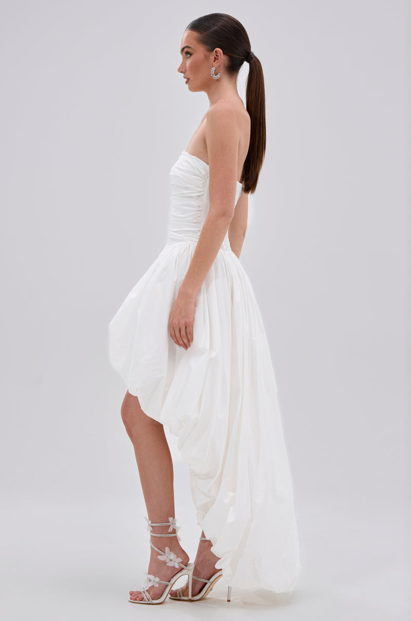 A woman stands in profile wearing the THIS AND THAT POPLIN HIGH LOW DRESS with a strapless bodice, voluminous skirt, and high-low hem. Her hair is styled in a sleek ponytail and she pairs the look with sheer floral-accented high-heeled sandals.