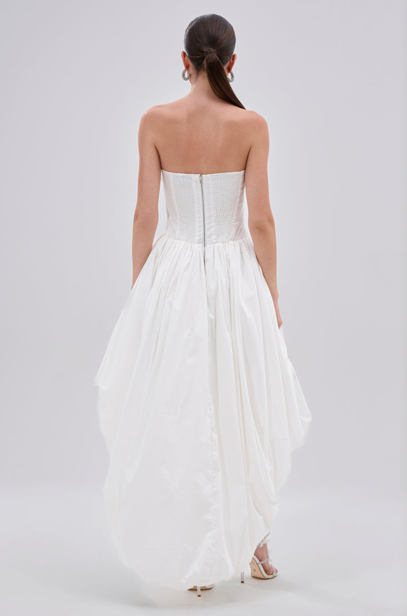 A woman with long dark hair in a ponytail is photographed from behind against a plain light background, wearing the THIS AND THAT POPLIN HIGH LOW DRESS featuring a strapless fitted bodice and voluminous skirt.