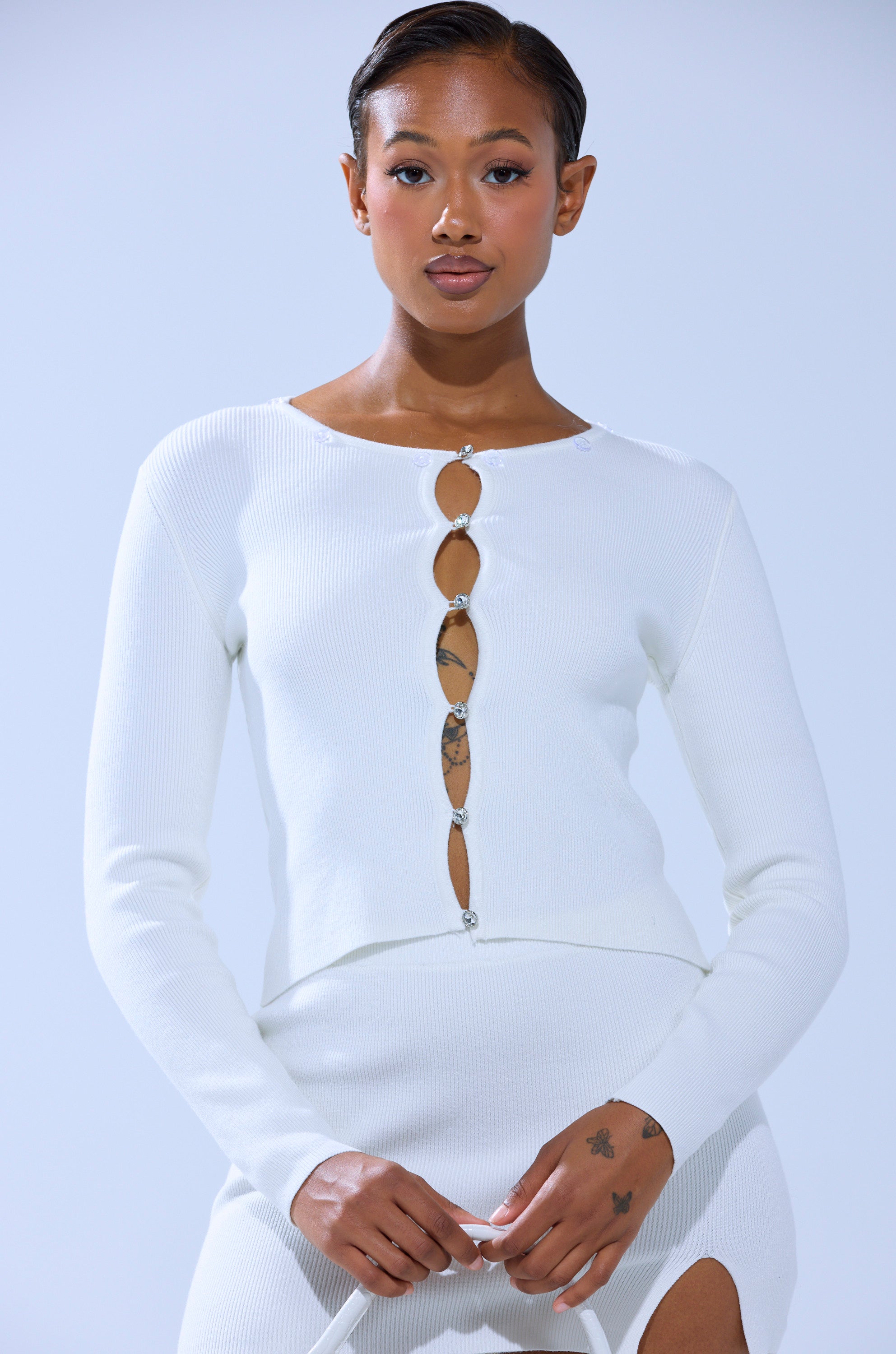 A person wearing the READY FOR THE SNOW CARDIGAN WITH FUR TRIM, a fitted white ribbed knit piece with decorative front clasps, stands against a light blue background, confidently looking at the camera.