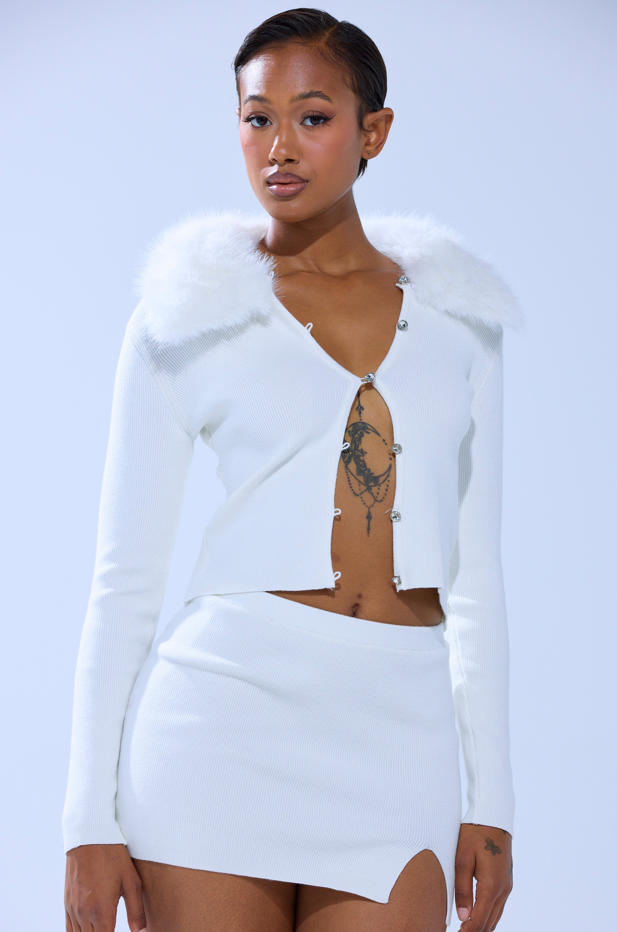 A woman with short hair and tattoos on her chest and hand wears the READY FOR THE SNOW CARDIGAN WITH FUR TRIM, featuring a white faux fur collar, silver clasps, and is paired with a matching mini skirt.