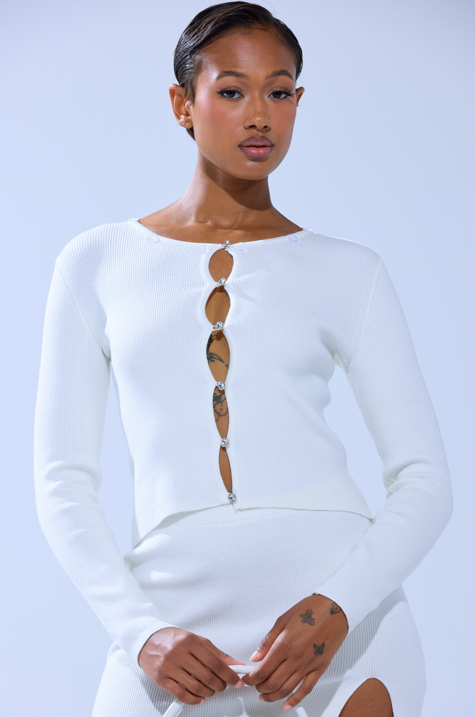 A woman with short, slicked-back hair models the READY FOR THE SNOW CARDIGAN WITH FUR TRIM and a matching skirt. Her tattooed hands and chest create a striking contrast against the light background.