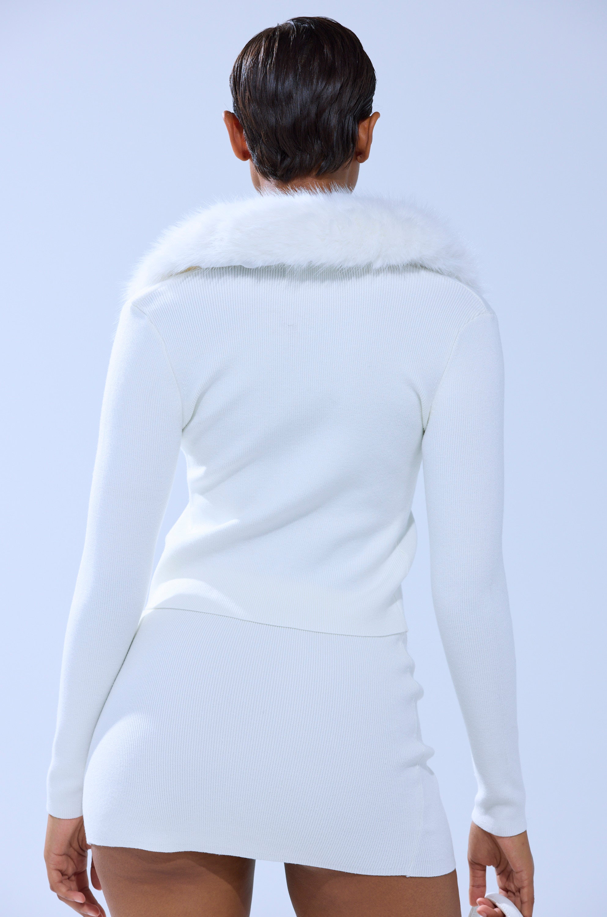 A person with short dark hair faces away, wearing the READY FOR THE SNOW CARDIGAN WITH FUR TRIM and a matching white mini skirt against a light blue background.