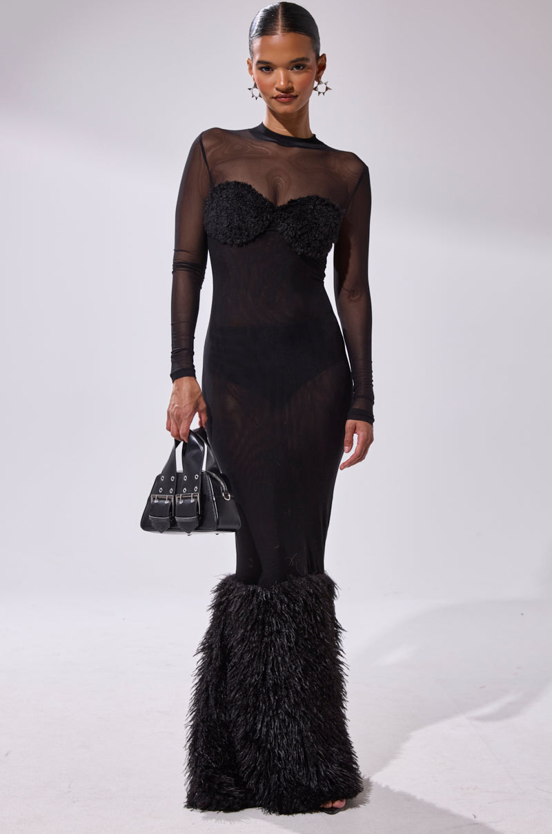 A woman poses against a plain light background wearing the RUNAWAY BRIDE MESH FUR MAXI DRESS, featuring a body-hugging fit, sheer black long sleeves, textured accents, and holding a black handbag.