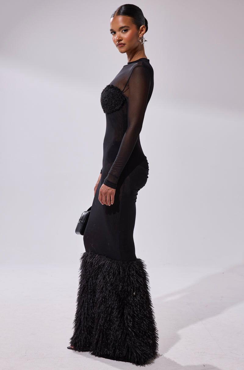 A woman models the RUNAWAY BRIDE MESH FUR MAXI DRESS, featuring a figure-hugging silhouette, sheer long sleeves, textured chest and back, and dramatic flared fur cuffs. She accessorizes with a small black bag against a plain light backdrop.