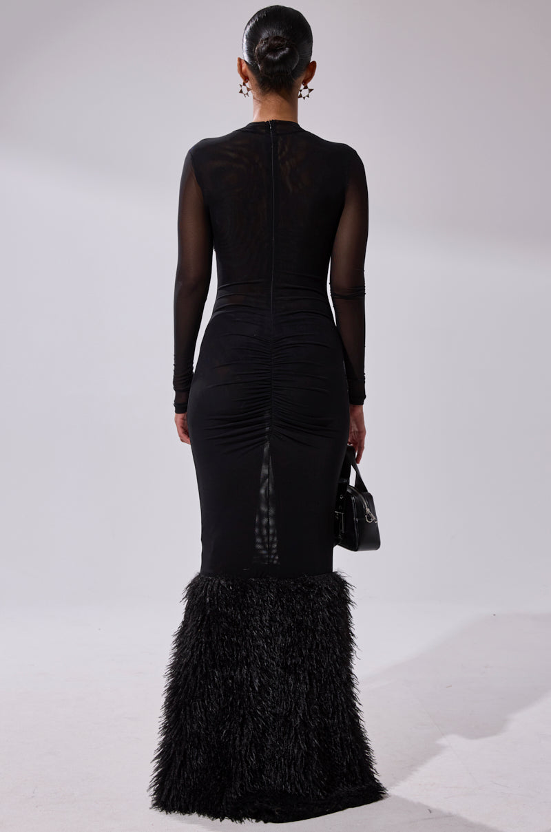 Someone faces away in the RUNAWAY BRIDE MESH FUR MAXI DRESS, a body-hugging black design with sheer mesh sleeves and faux fur trim at the hem. They hold a small handbag and wear their hair in an updo against a plain light backdrop.