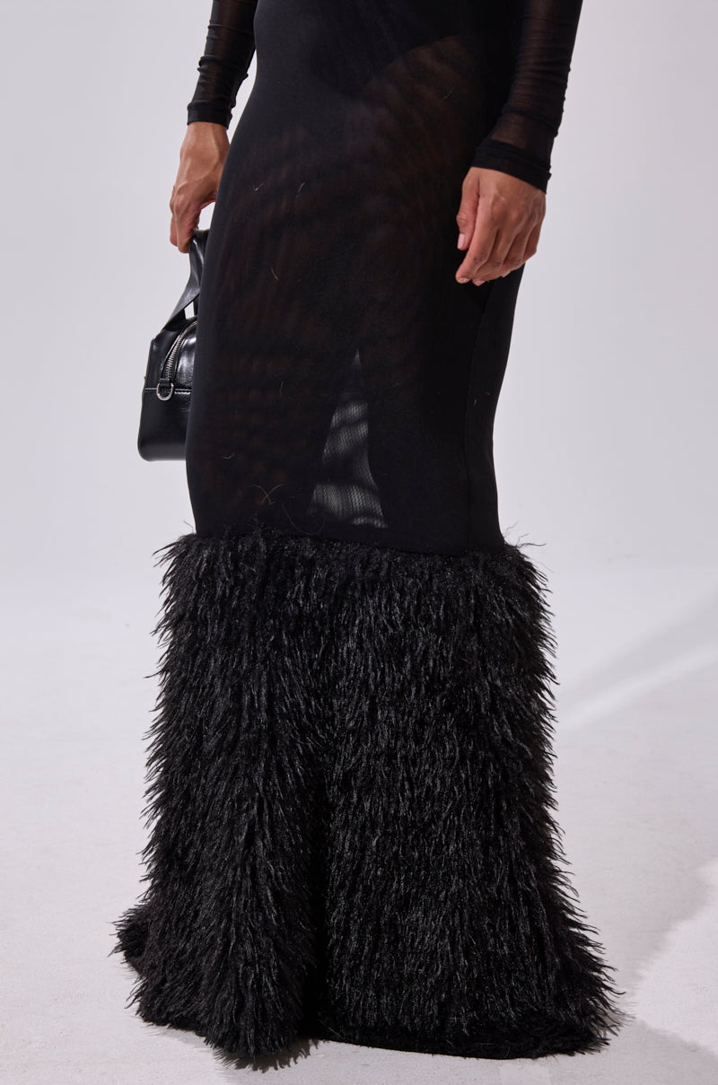 A person is wearing the RUNAWAY BRIDE MESH FUR MAXI DRESS, which features a body-hugging fit, sheer fabric, and fluffy textured black fur at the skirt's hem, holding a small black handbag against a plain light background.