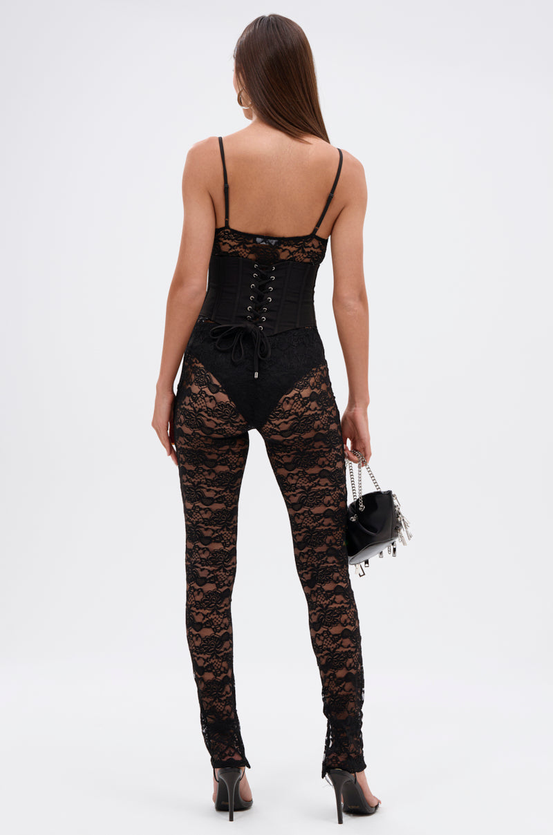 A woman faces away, wearing the ACTING BRAND NEW LACE JUMPSUIT WITH CORSET BELT and high heels. She holds a small black handbag with silver details in her left hand against a plain white background.