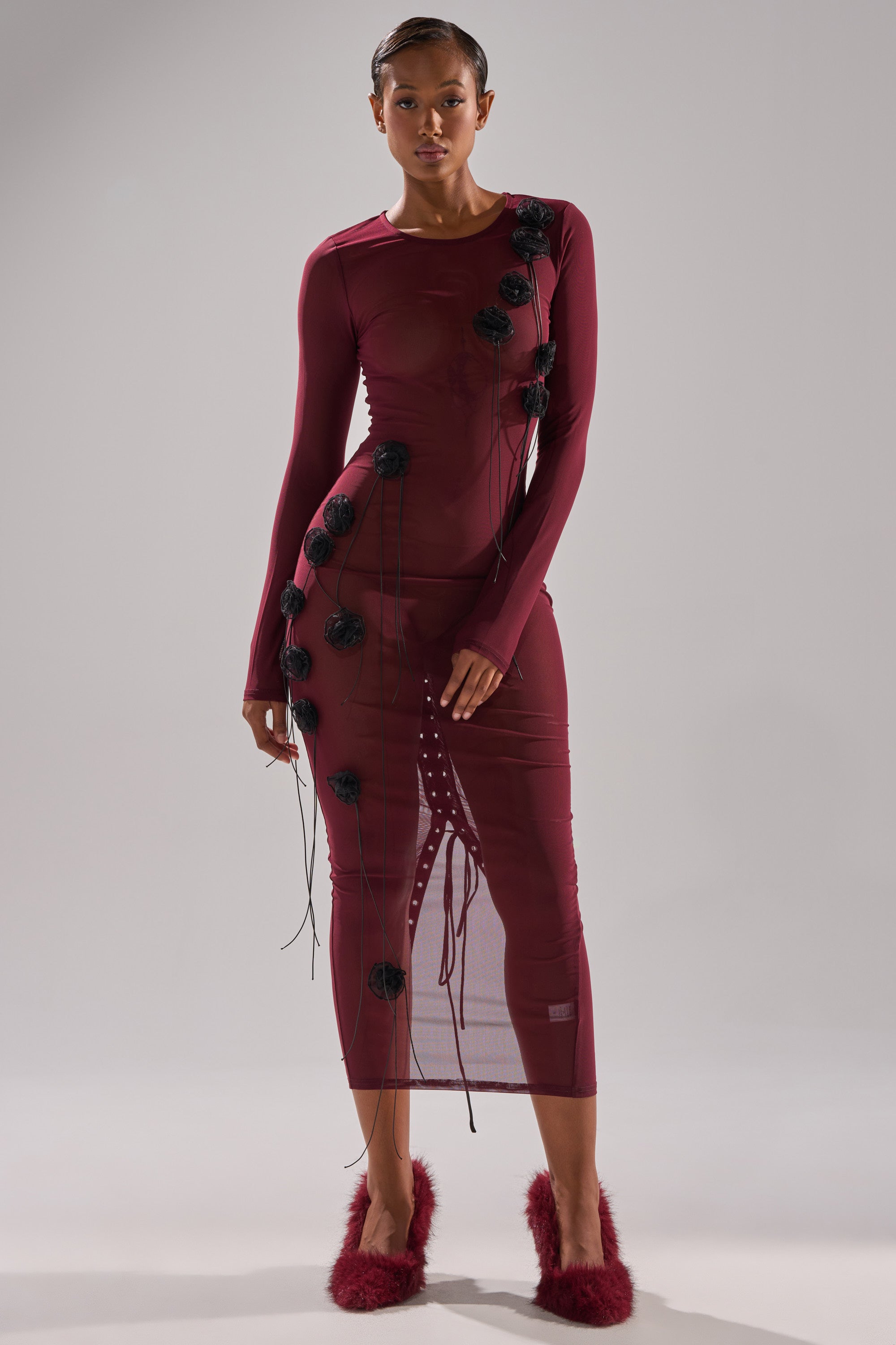 A woman wears the OBVIOUSLY MESH MIDI DRESS IN BURGUNDY, featuring a fitted silhouette with black floral details and side strings, paired with burgundy fuzzy slippers against a plain light gray background.