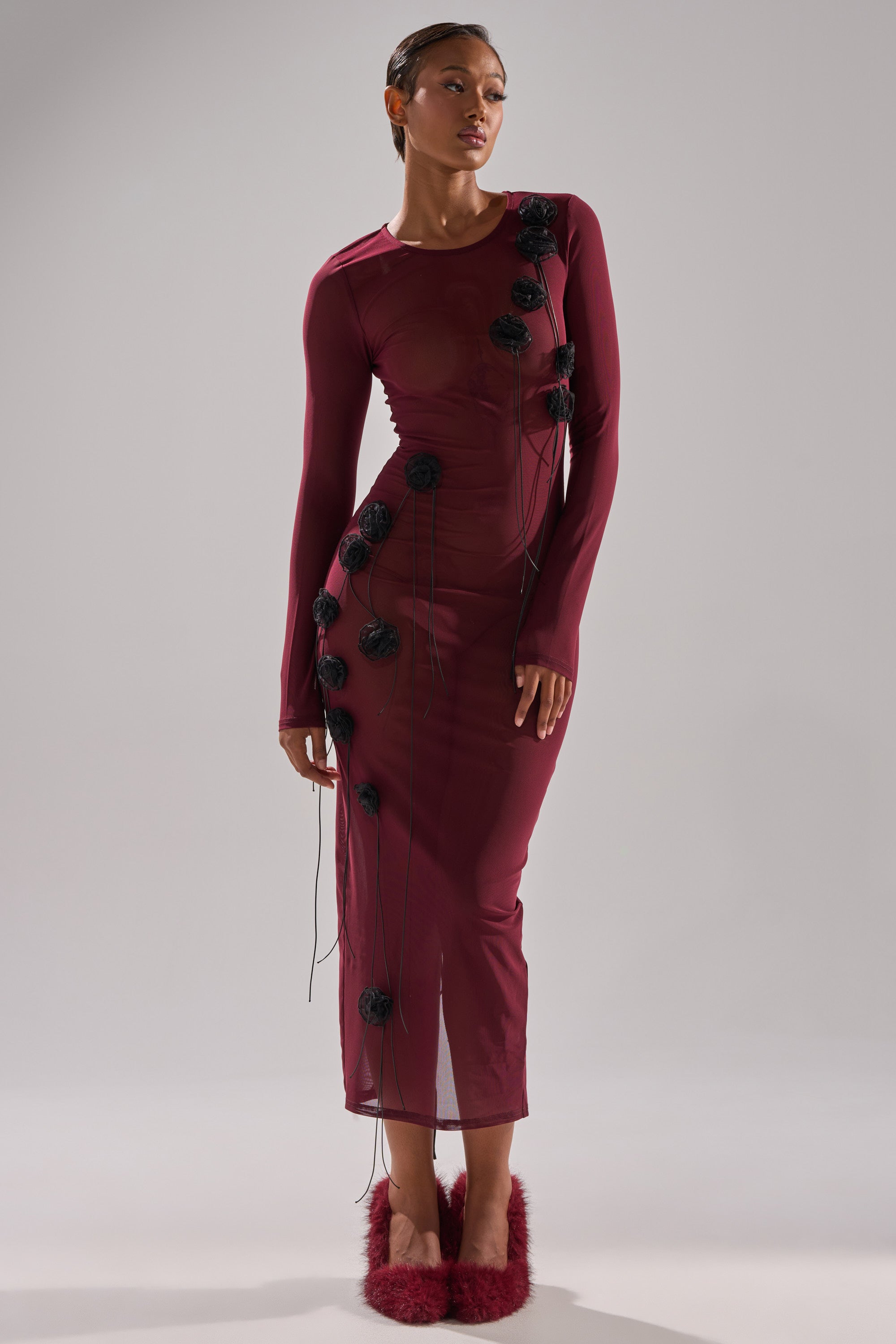 A woman wears the OBVIOUSLY MESH MIDI DRESS IN BURGUNDY, a sheer, long-sleeved mesh midi dress with black floral appliqués and strings, featuring a lace-up back. She pairs it with burgundy fluffy shoes against a plain gray background.