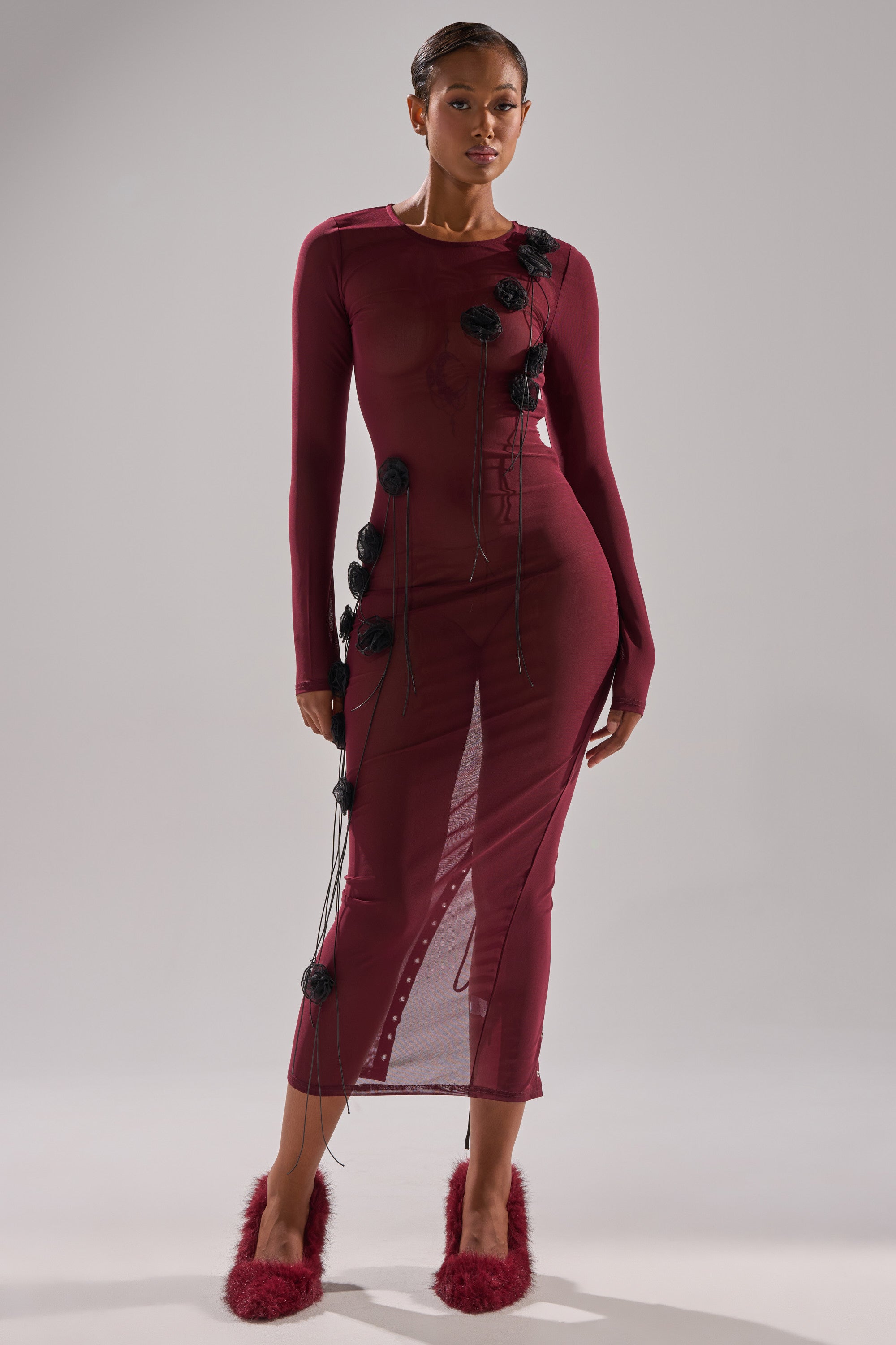 A woman poses in the OBVIOUSLY MESH MIDI DRESS IN BURGUNDY—a fitted, long-sleeve mesh midi dress with black floral appliqués—paired with burgundy fuzzy-heeled shoes, against a light gray background.