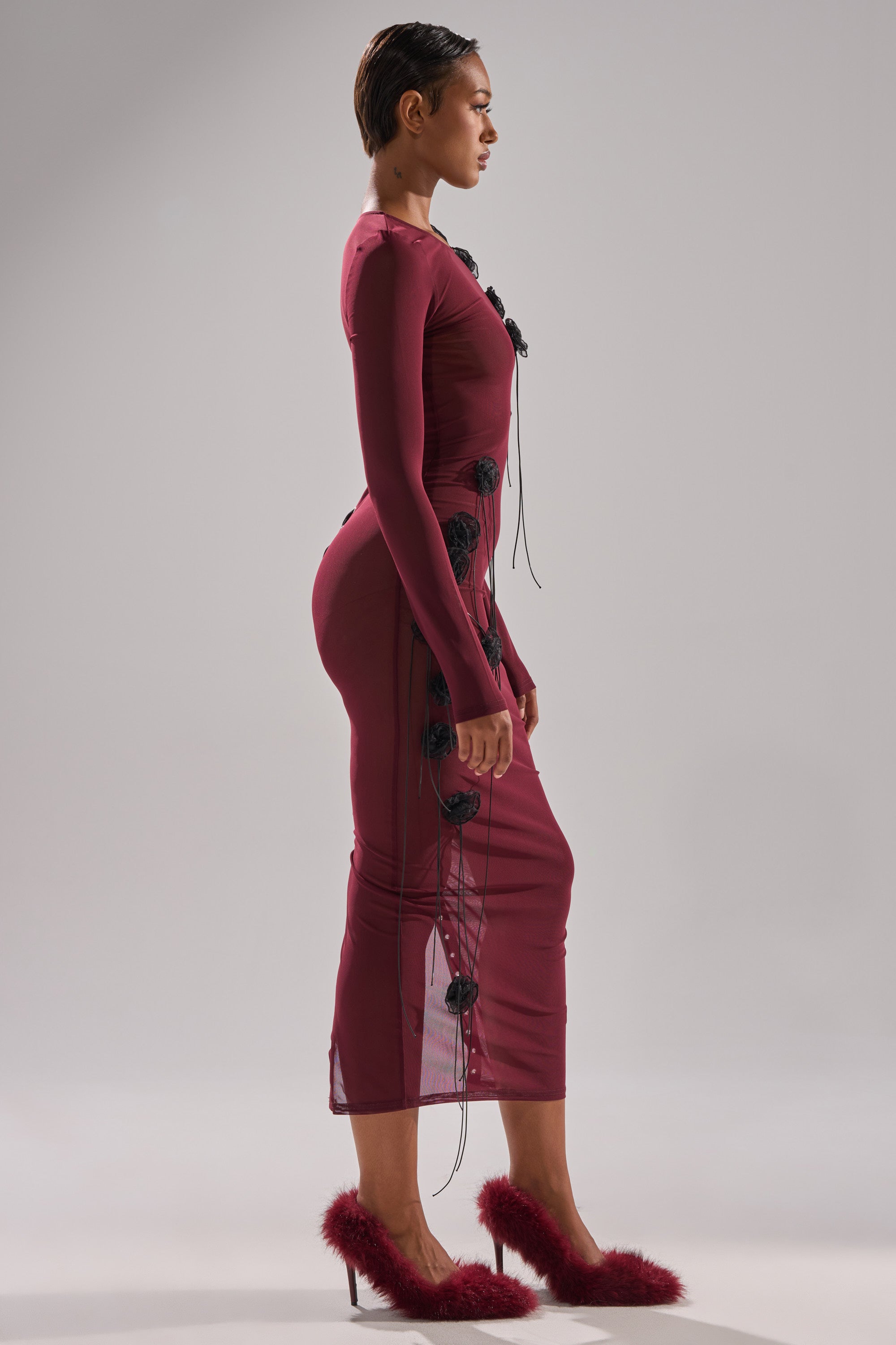 A woman in profile wears the OBVIOUSLY MESH MIDI DRESS IN BURGUNDY—a fitted, sheer dress with black accents—paired with burgundy fluffy high heels, posing against a plain light backdrop.