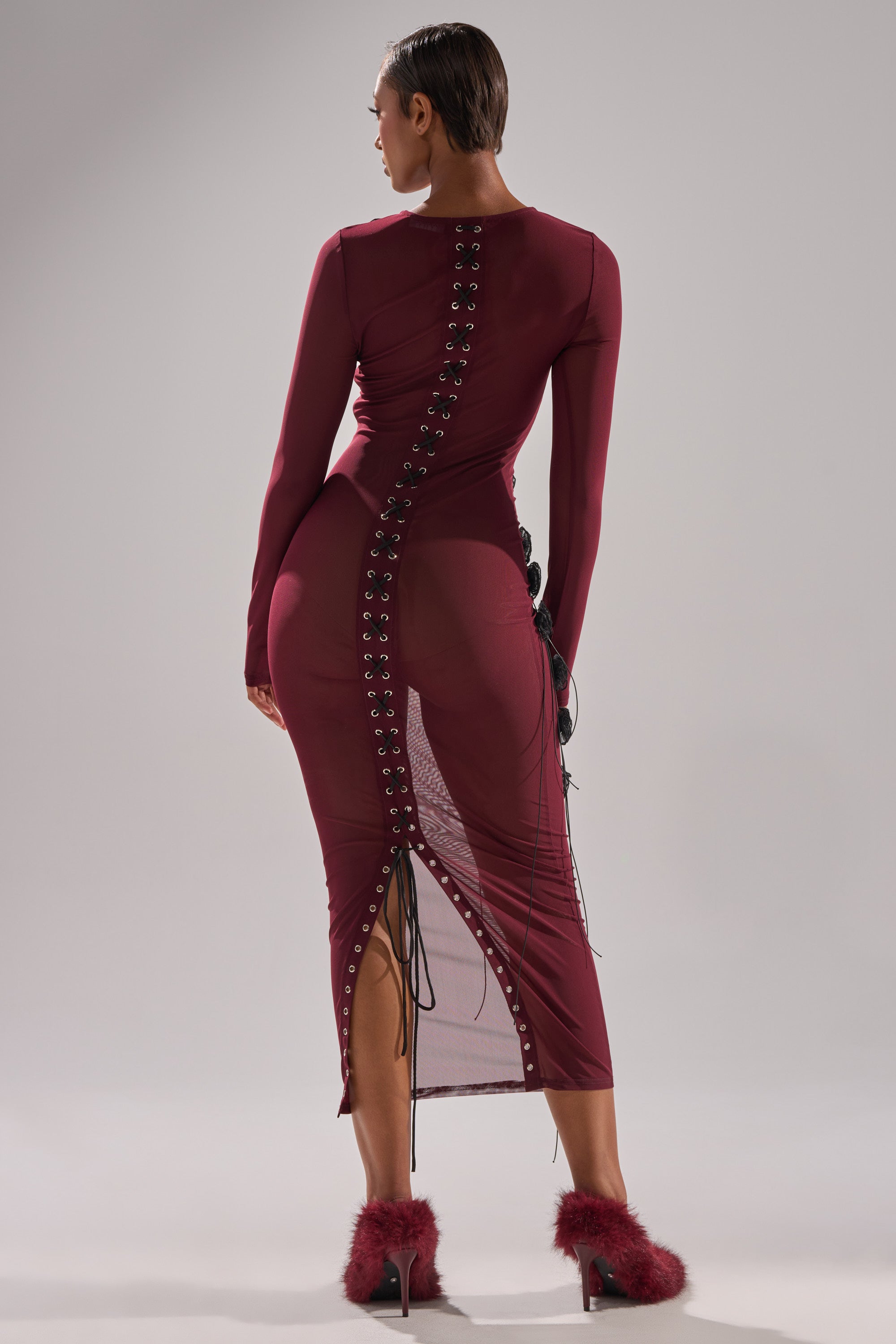 A person faces away, wearing the OBVIOUSLY MESH MIDI DRESS IN BURGUNDY—fitted with lace-up grommet back and sheer mesh panel—paired with burgundy fluffy heels against a plain light gray backdrop.