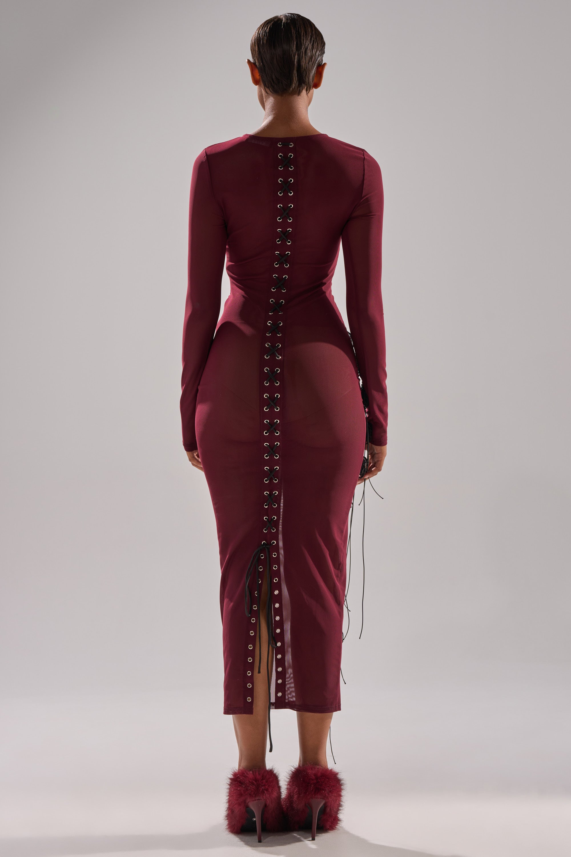 A person stands facing away, wearing the OBVIOUSLY MESH MIDI DRESS IN BURGUNDY, a fitted long-sleeved dress with a lace-up back and grommet detailing, paired with matching burgundy high heels featuring feathery accents.
