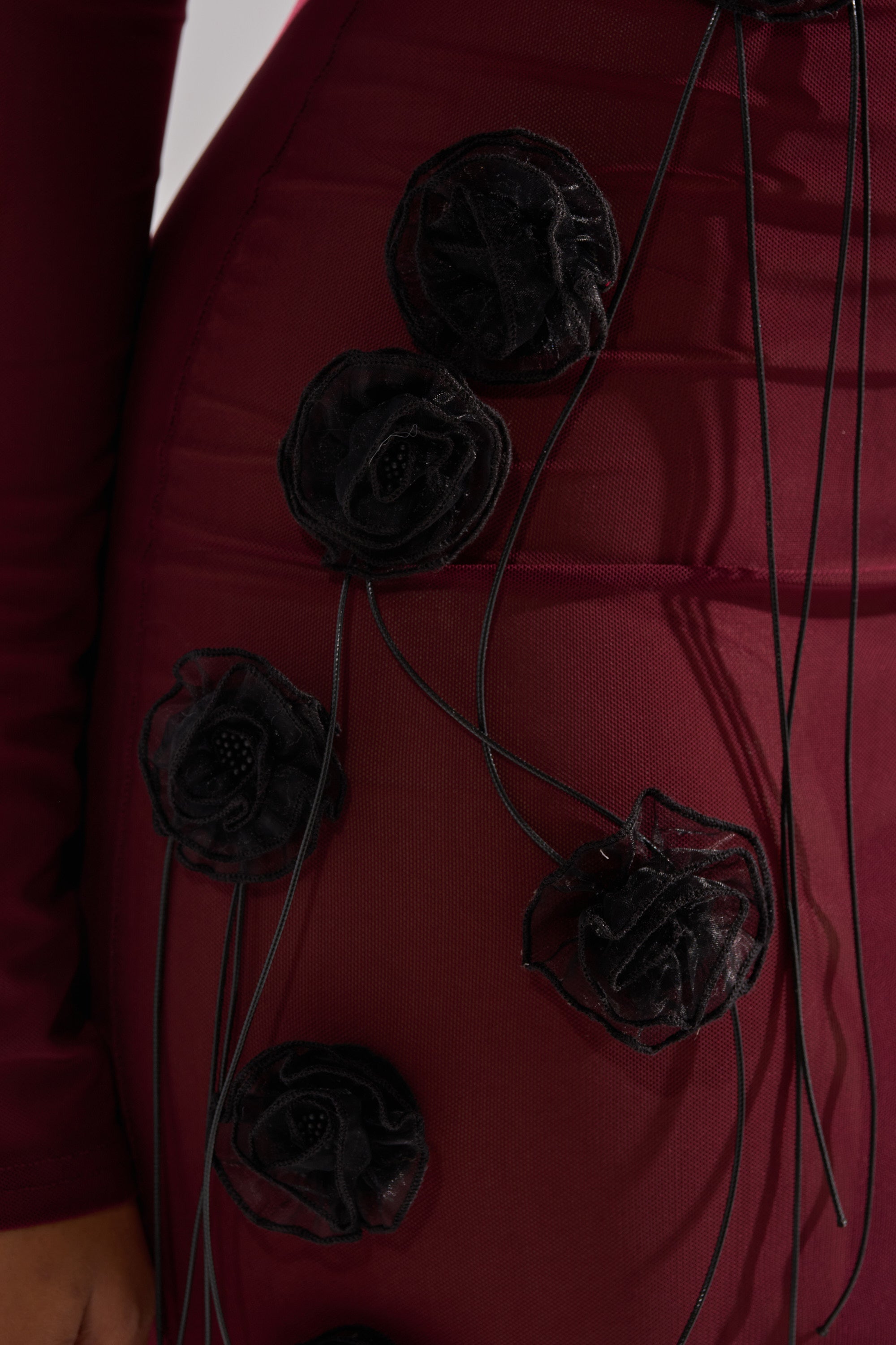 A close-up of the OBVIOUSLY MESH MIDI DRESS IN BURGUNDY, featuring black fabric rosettes and thin black cords, highlights part of a person’s arm and hip.