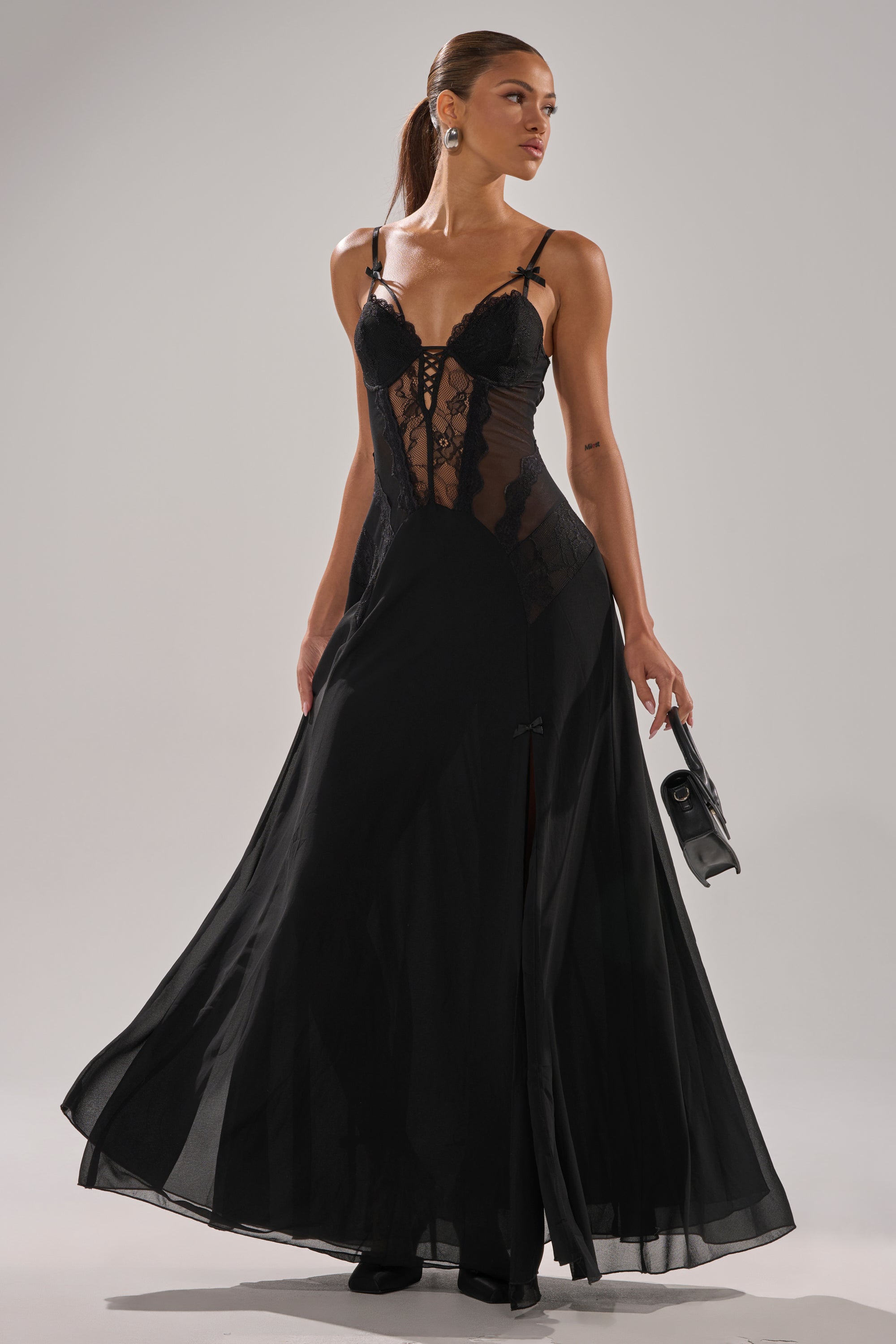 Wearing the LEILA MAXI DRESS—a sheer black lace bustier gown with a fitted bodice, spaghetti straps, and floor-length silhouette—a woman stands against a plain background, holding a black clutch and sporting a sleek ponytail.