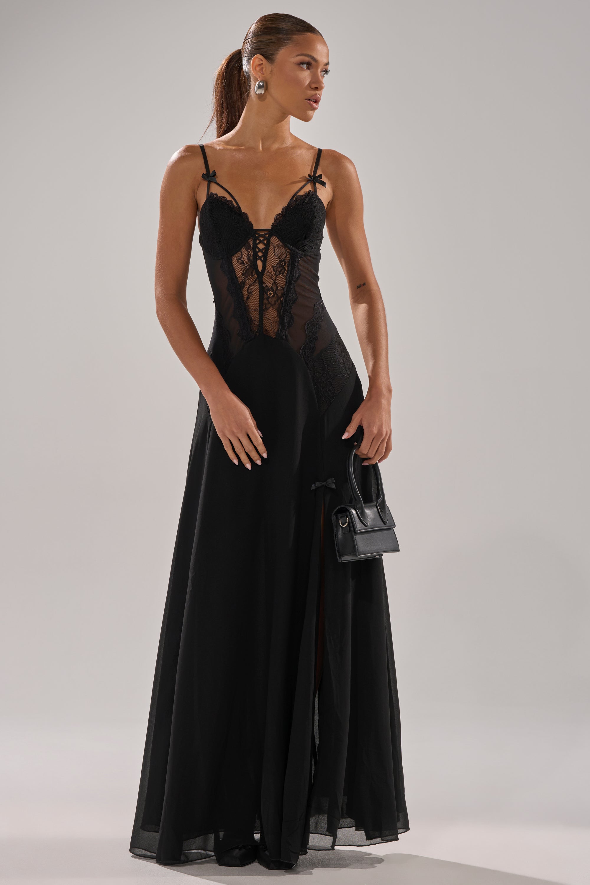 A woman in the LEILA MAXI DRESS, with a sheer lace bodice and thigh-high slit, holds a small black handbag. Her hair is tied back as she gazes to the side against a plain background.