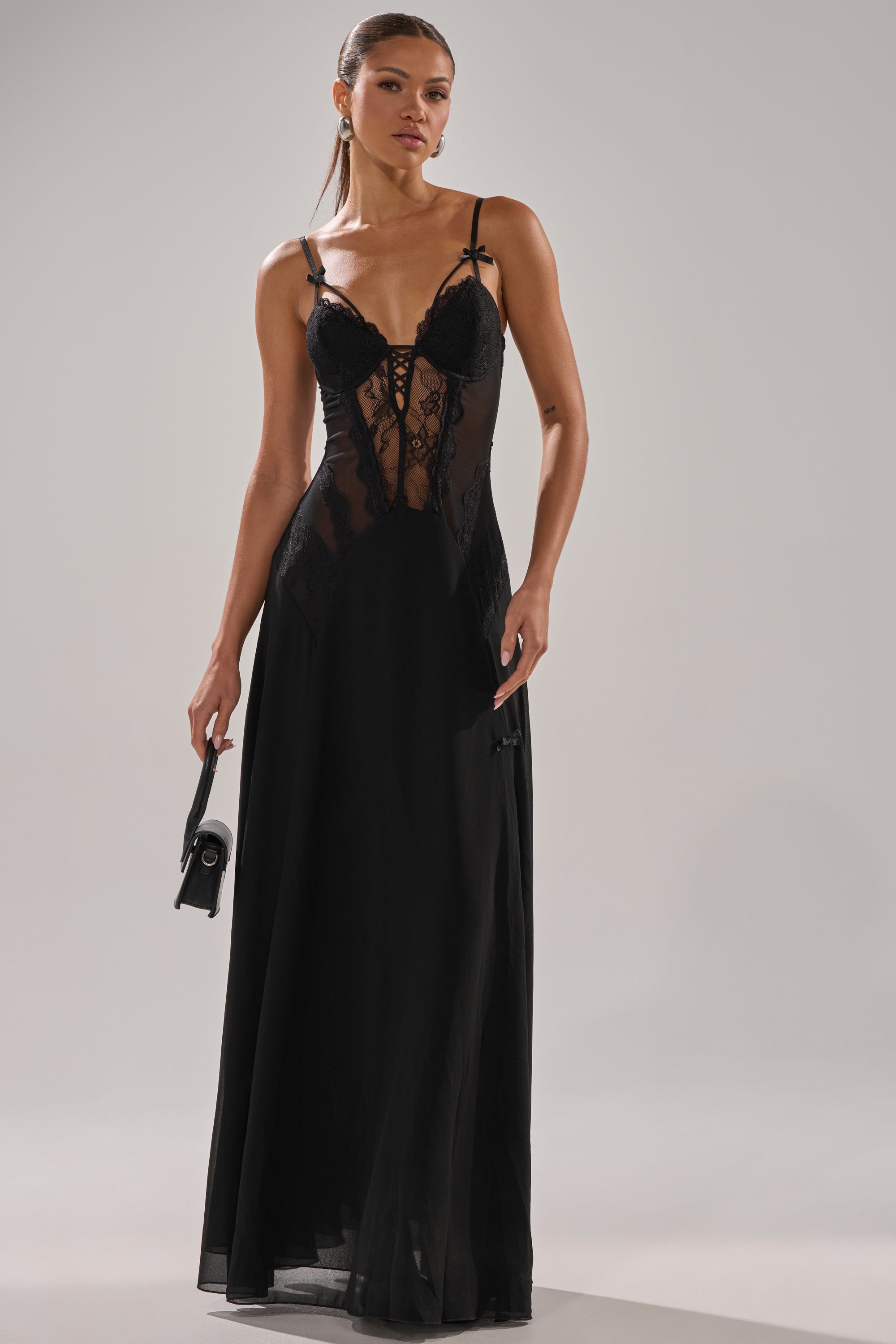 A woman wearing the LEILA MAXI DRESS—a long black lace bustier dress with spaghetti straps and a sheer bodice—holds a small black handbag and stands against a plain light background.