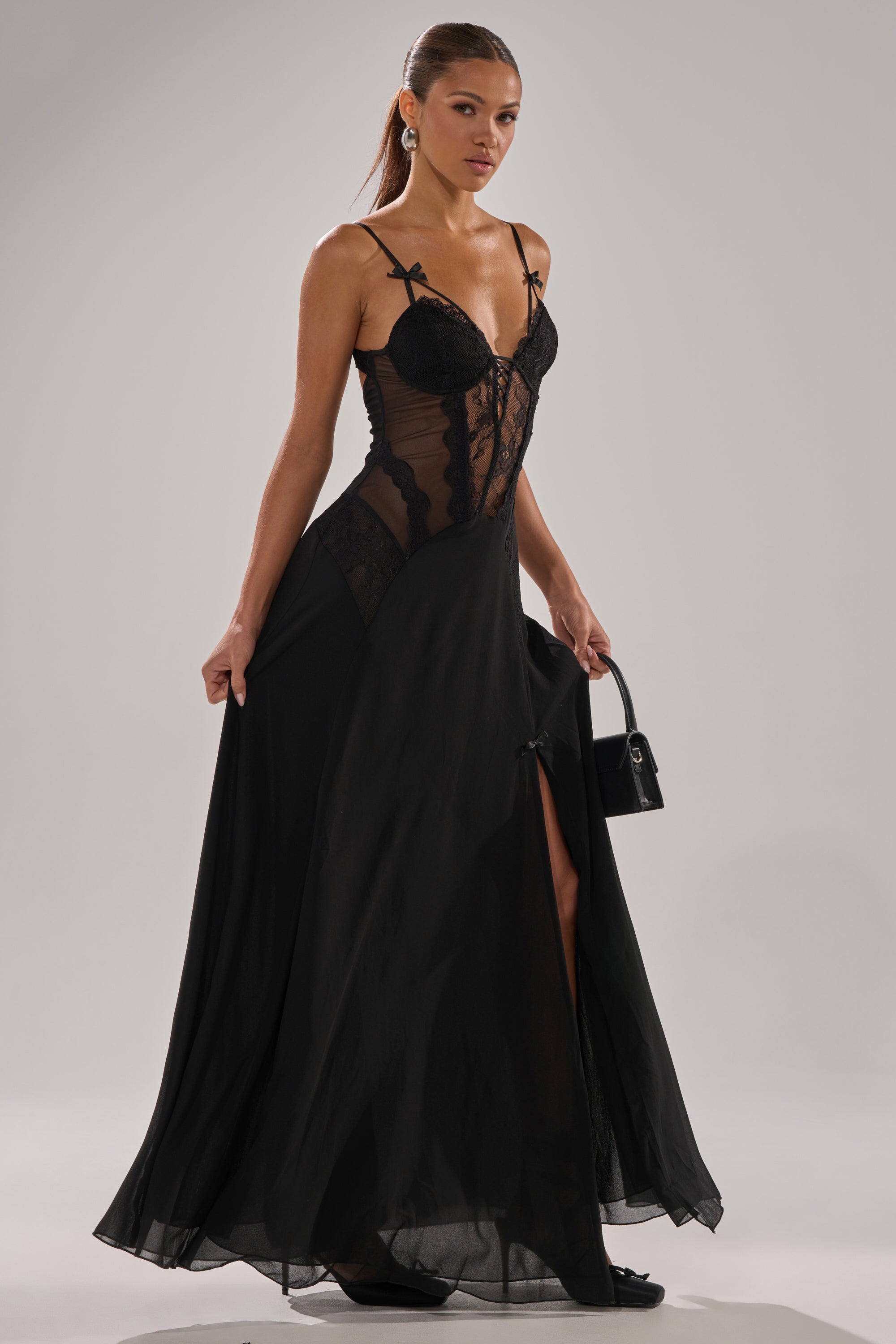 A woman poses confidently in the LEILA MAXI DRESS, a sheer black lace bustier gown with a floor-sweeping silhouette, paired with a small black handbag and statement earrings.