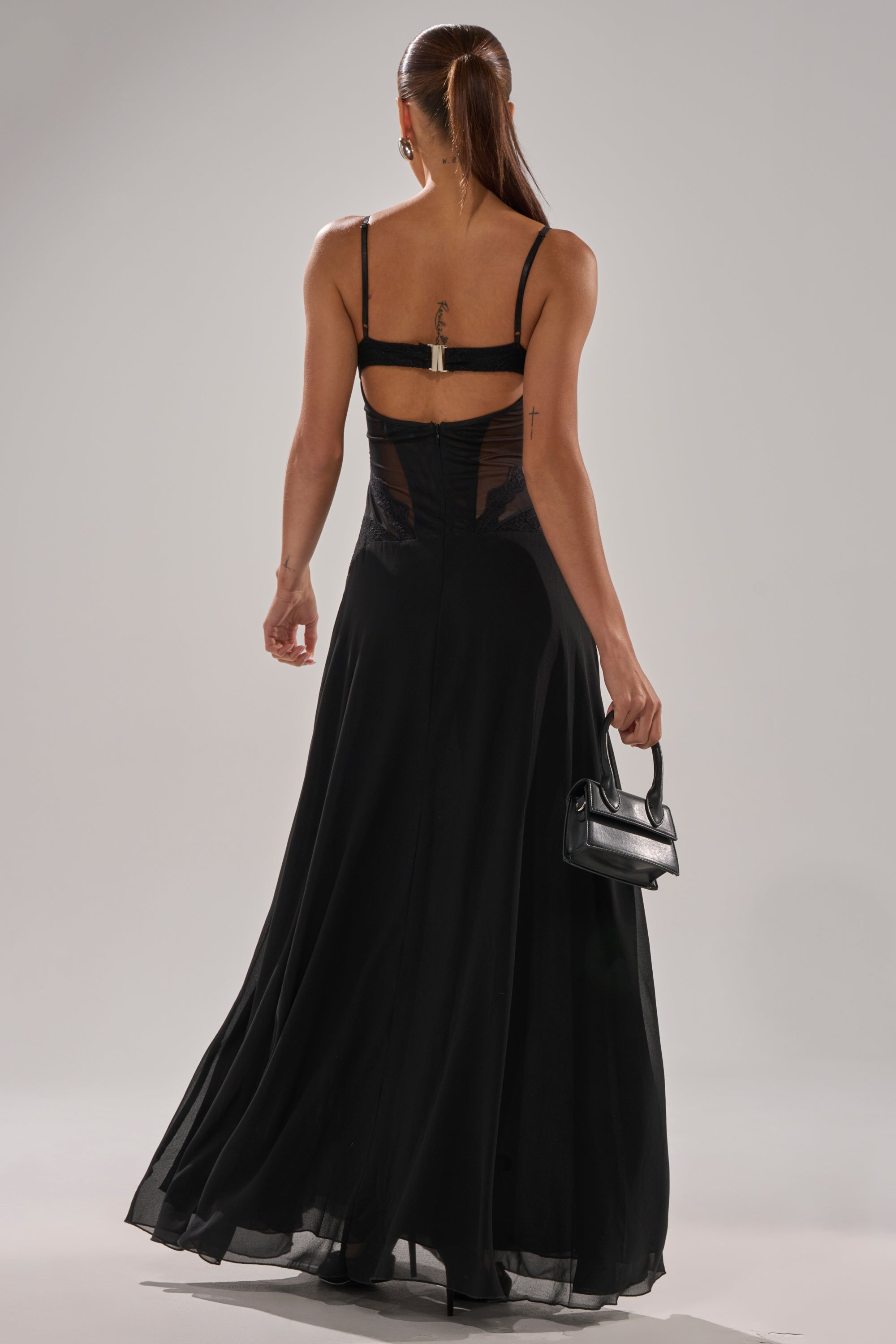 A woman with long brown hair in a ponytail stands with her back to the camera, wearing the LEILA MAXI DRESS—a sleeveless, floor-length black dress with sheer panels—and holding a small black handbag.