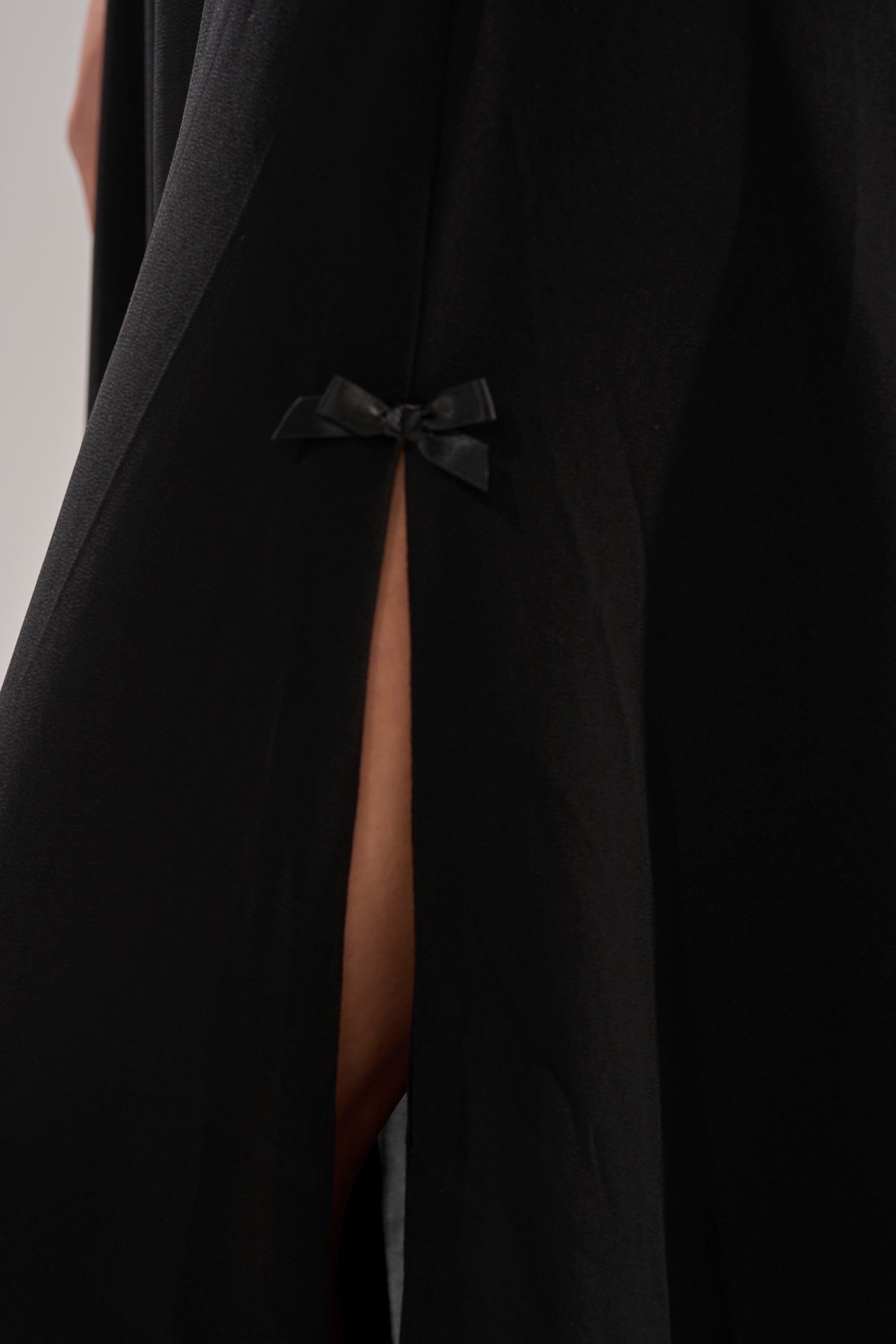 A close-up of the LEILA MAXI DRESS, a black thigh-high slit dress adorned with a small black bow and revealing a hint of skin through the opening.