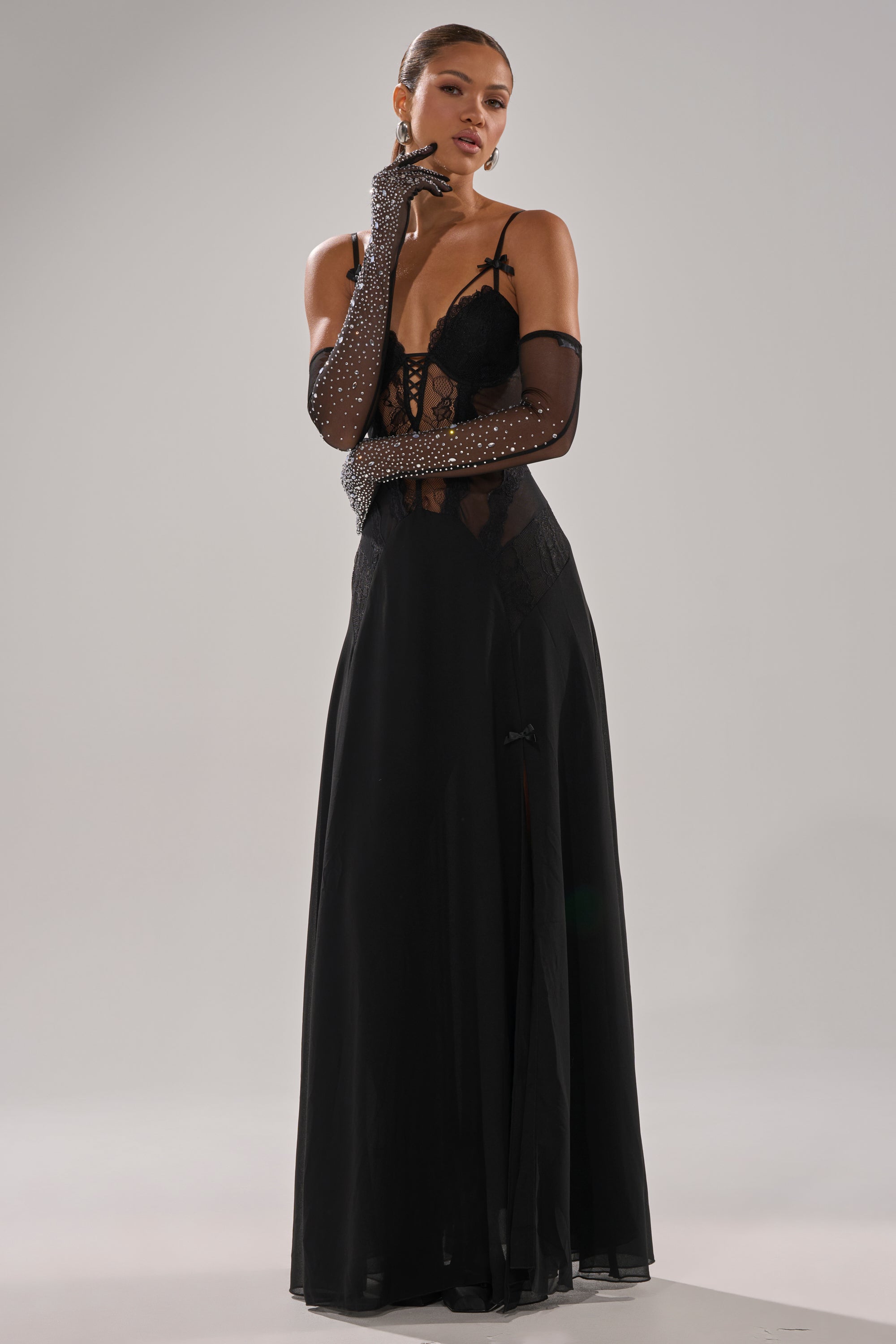 A woman poses confidently in the LEILA MAXI DRESS, a long, elegant lace bustier style. She accessorizes with sheer, elbow-length gloves adorned with sparkling stones and wears her hair pulled back against a light background.