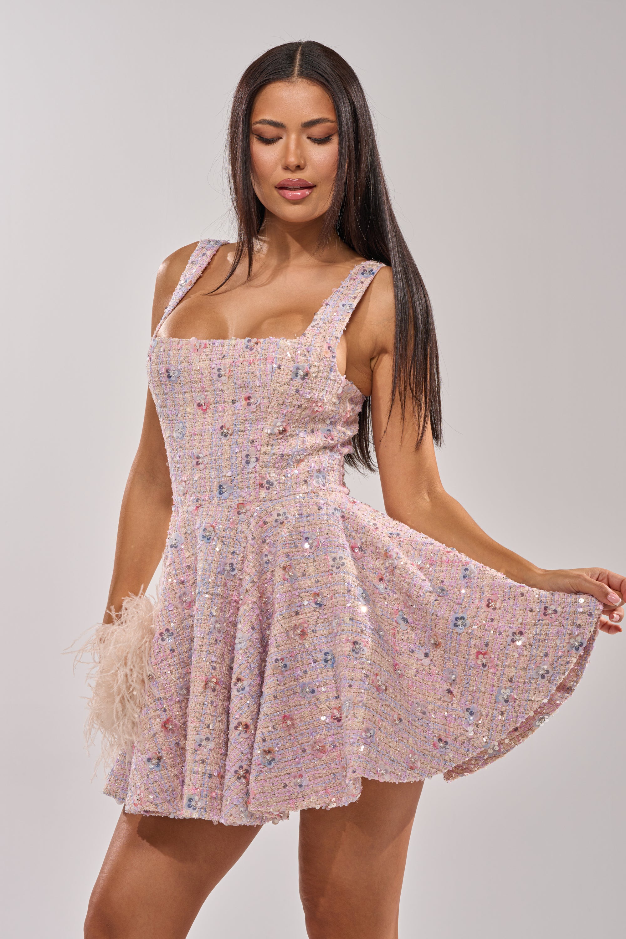 A woman with long, straight dark hair wears the SWEET LIKE CANDY MINI DRESS—a sleeveless, square-neck pastel mini dress with multicolored embellishments—holding out the skirt against a plain light background. 0