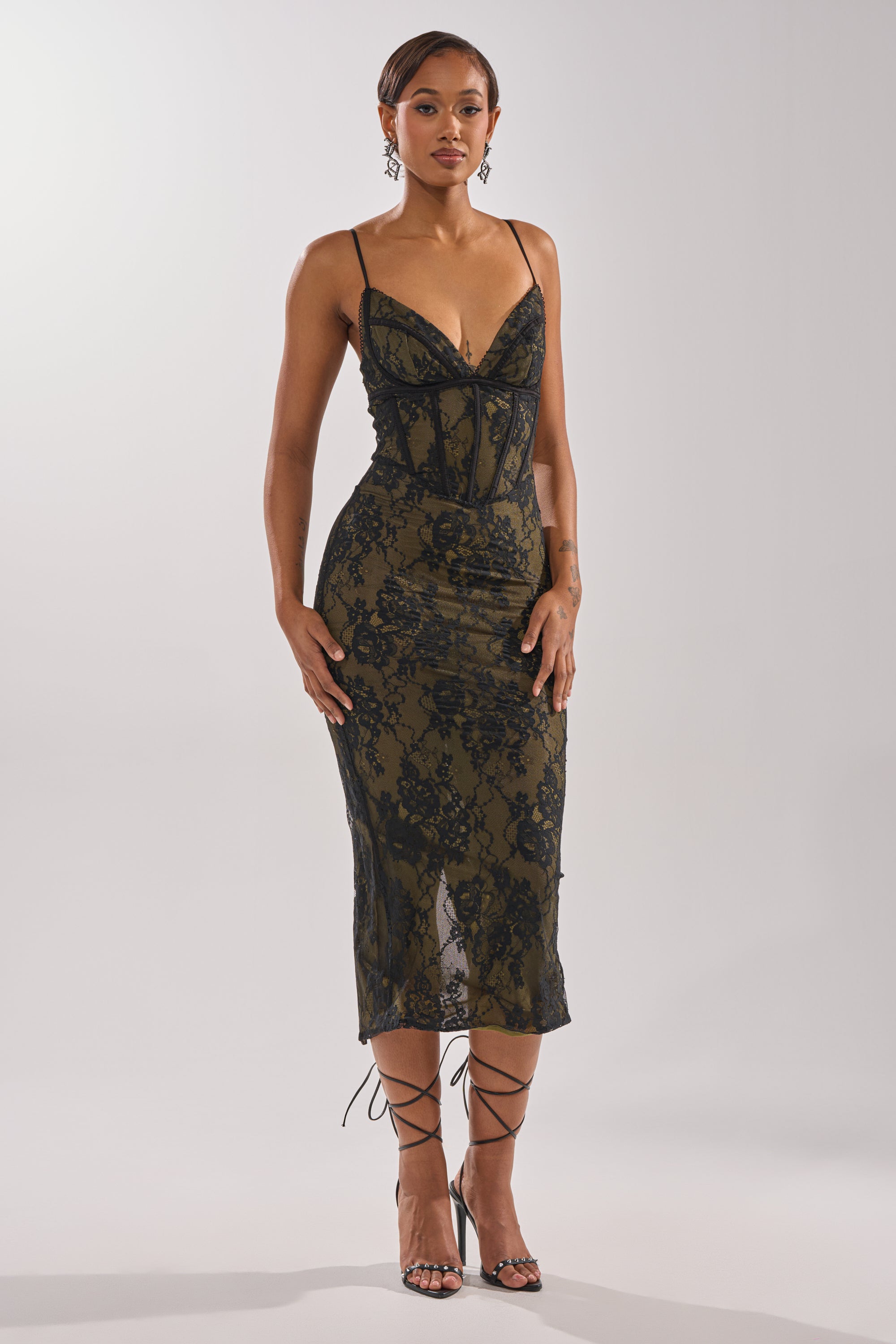 A woman poses against a light background in the BETTER BELIEVE IT LACE MAXI DRESS—a fitted, sleeveless black lace gown with thin straps and a deep neckline—styled with short hair, dangling earrings, and strappy high heels for an elegant evening look. 1