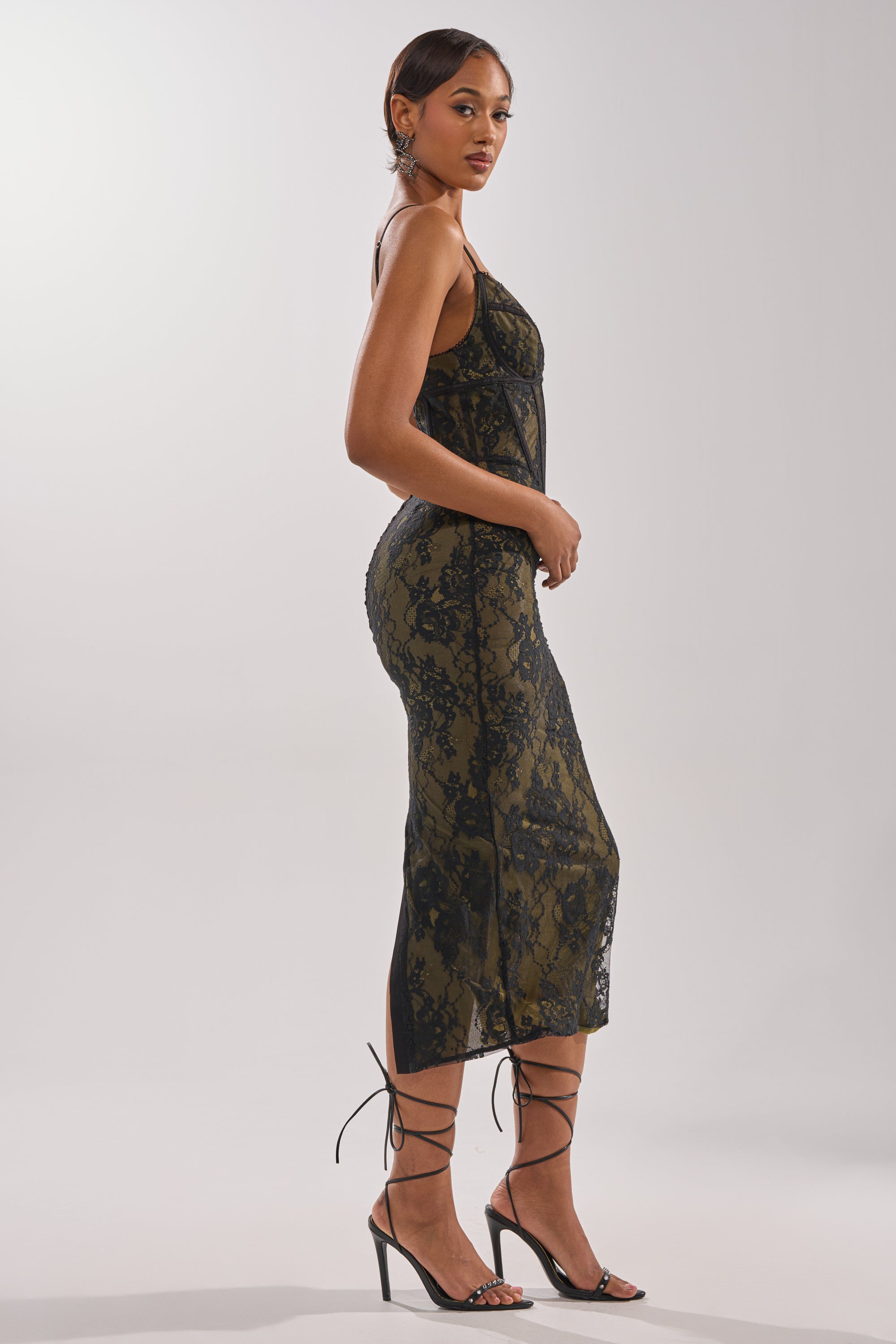 A woman stands sideways in the BETTER BELIEVE IT LACE MAXI DRESS—a sleeveless, form-fitting maxi dress with a corset bodice—paired with strappy black heels against a light neutral background. 3