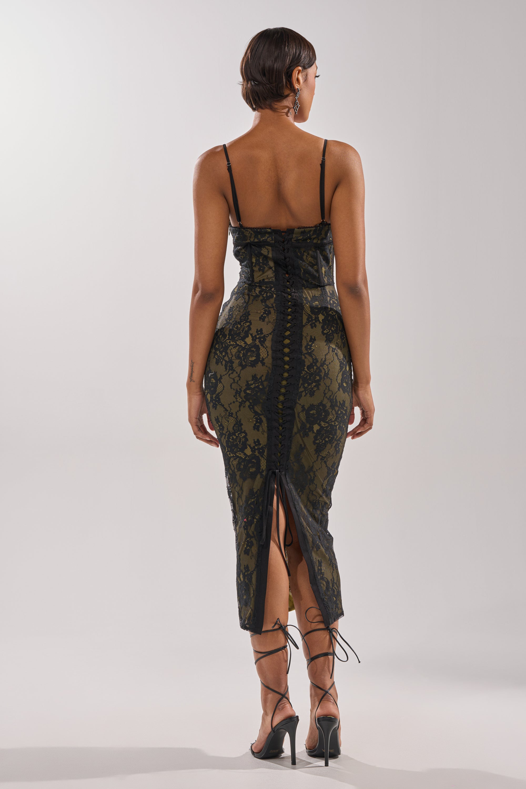 A person models the BETTER BELIEVE IT LACE MAXI DRESS, a form-fitting, dark green maxi with spaghetti straps, corset bodice, lace-up back, and intricate lace detailing, paired with high-heeled strappy sandals. 4