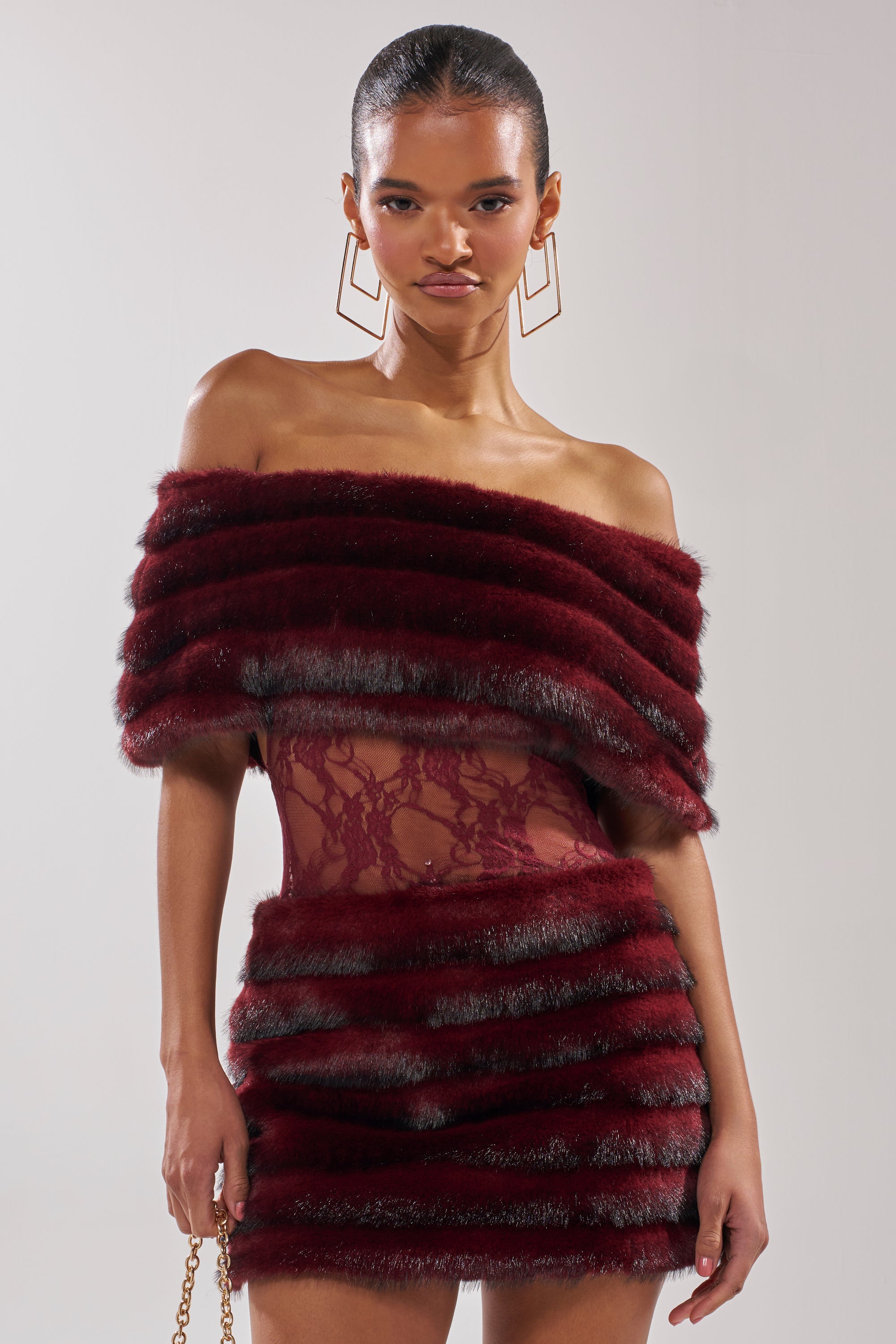 A woman wears the SOFT CHERRY SLEEVELESS MINI DRESS with faux-fur panels and sheer lace waist detail, styled with geometric gold earrings, posing against a plain light background.