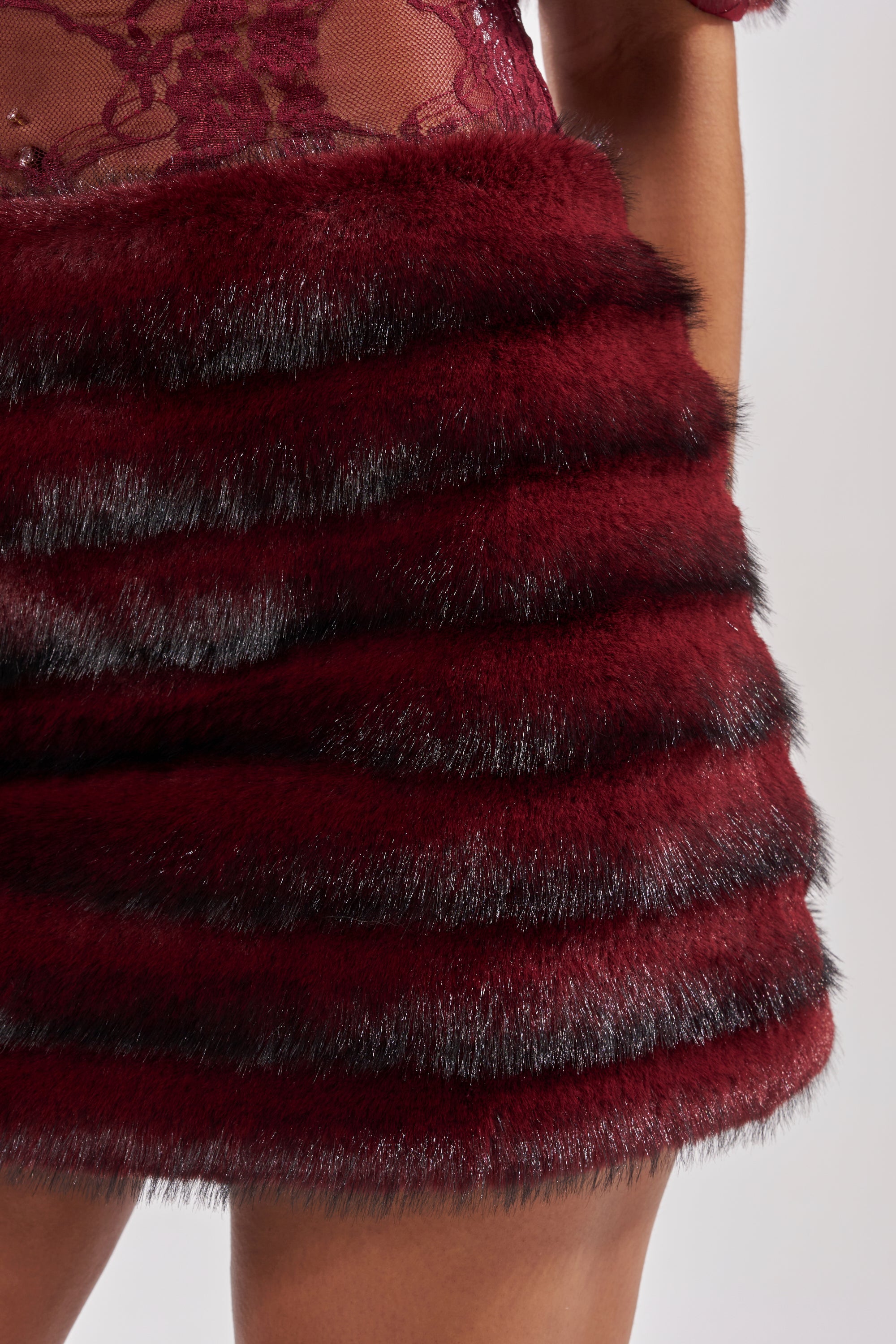A close-up shows someone wearing the SOFT CHERRY SLEEVELESS MINI DRESS in deep red with black-striped, textured faux-fur tiers; part of their arm and a sheer lace top with delicate detailing is visible.