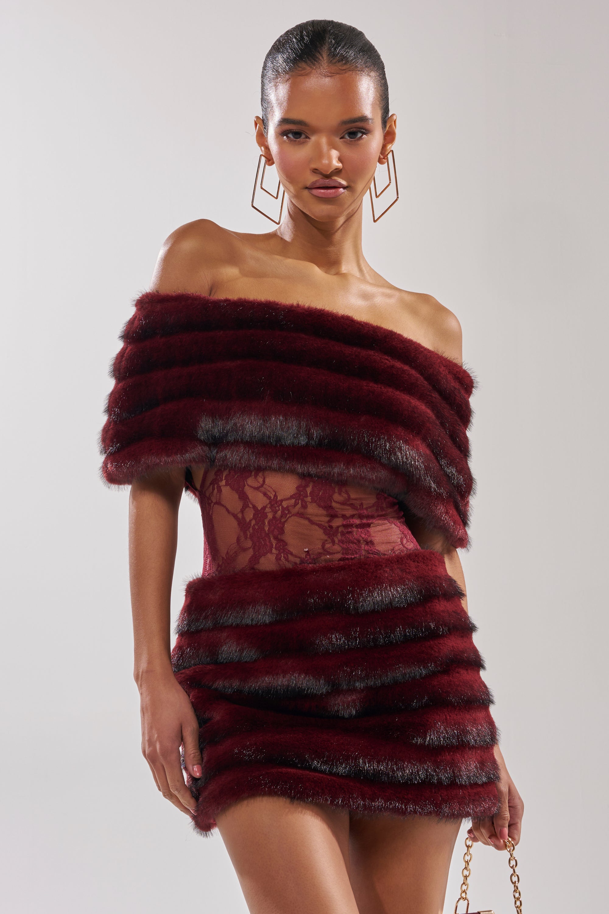 A woman in the SOFT CHERRY SLEEVELESS MINI DRESS with horizontal faux fur bands, off-the-shoulder capelet styling, and sheer lace details accessorizes with geometric earrings and a gold chain purse against a plain background.