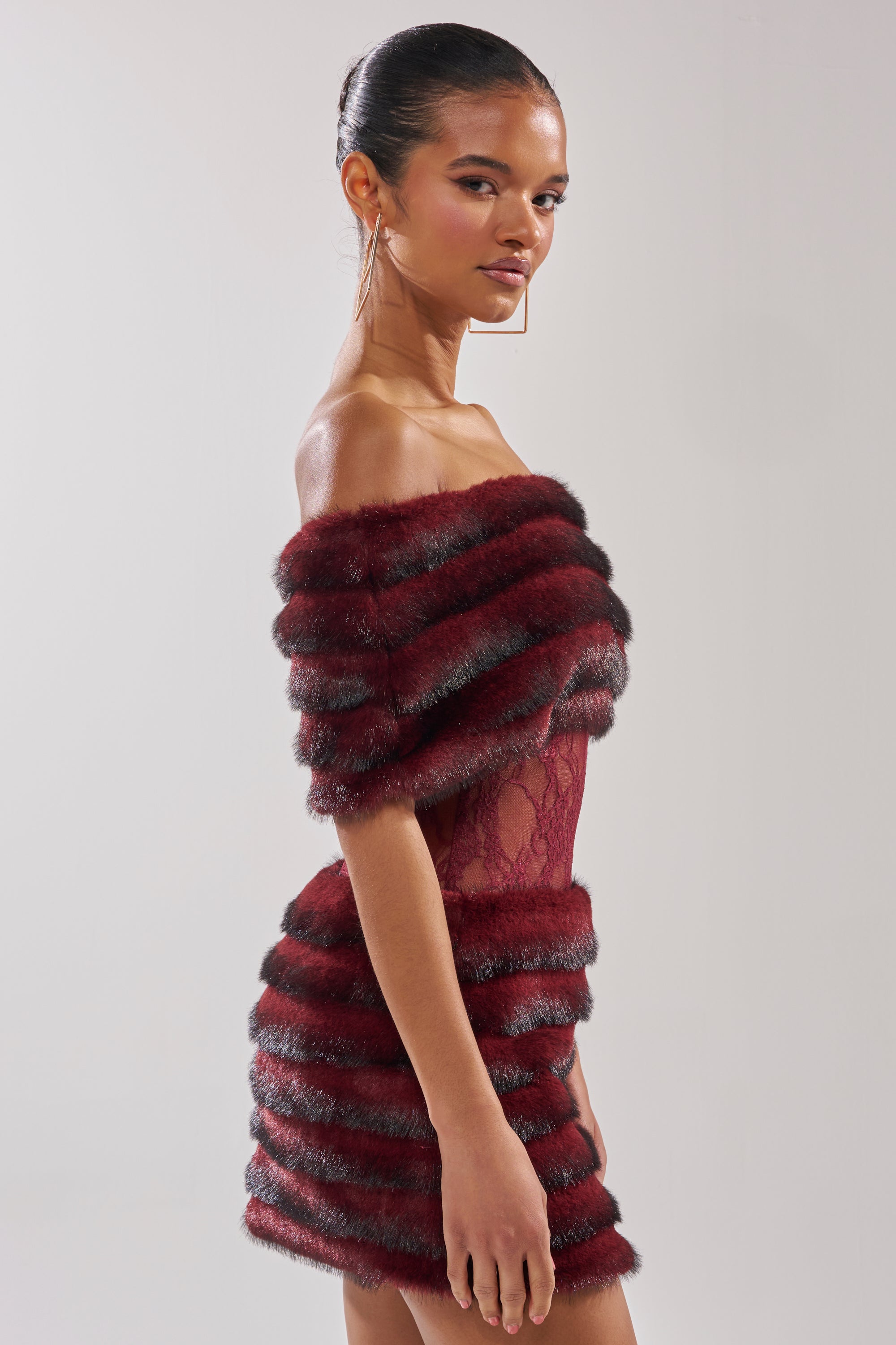 A woman poses in the SOFT CHERRY SLEEVELESS MINI DRESS, featuring burgundy lace, horizontal fur stripes, and sheer details. She accessorizes with large hoop earrings and wears her hair in a sleek bun.