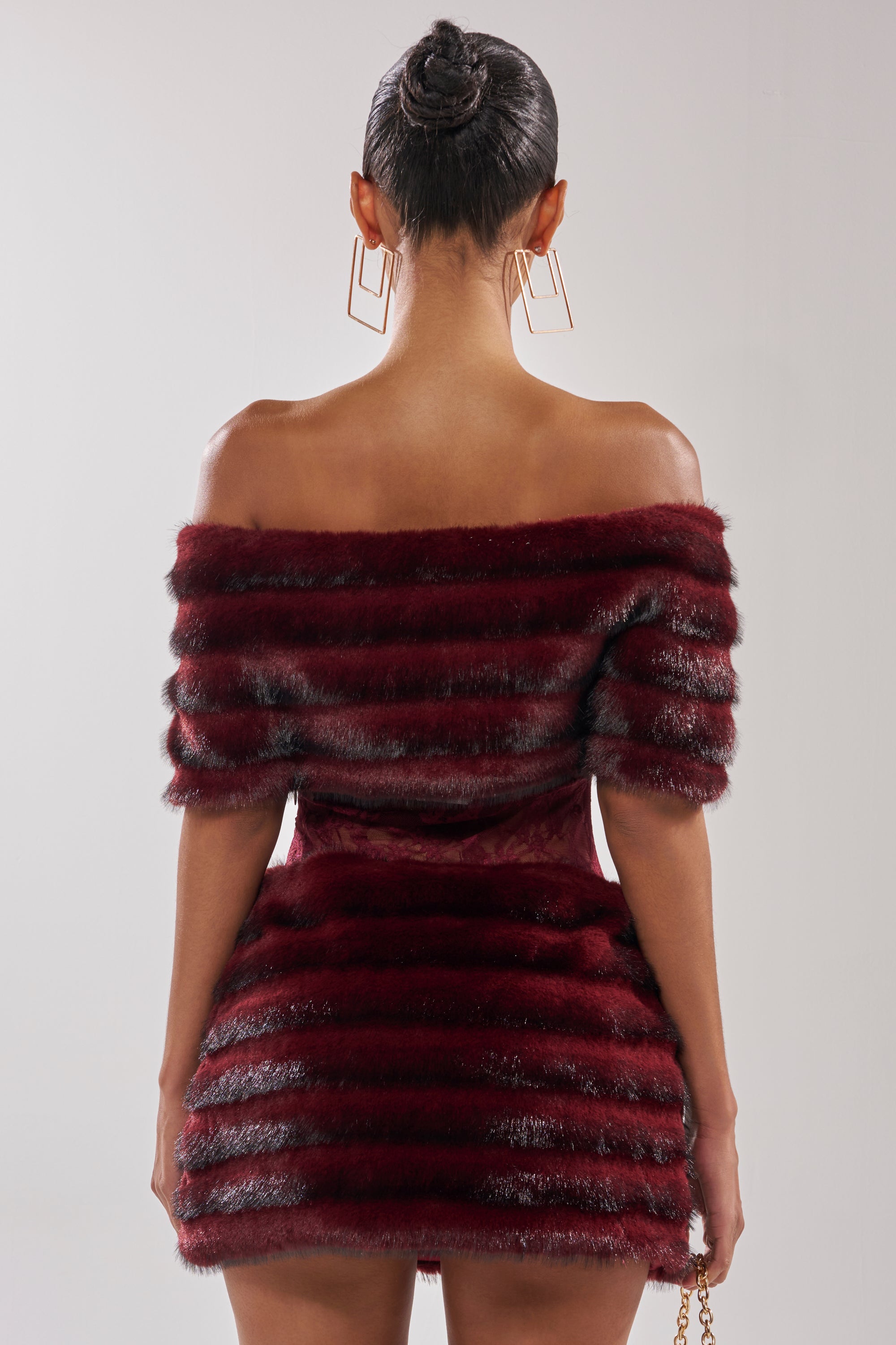 A woman faces away from the camera in the SOFT CHERRY SLEEVELESS MINI DRESS, featuring burgundy fabric with black striped faux-fur tiered panels. Her hair is styled in a sleek bun, and she accessorizes with bold geometric earrings.