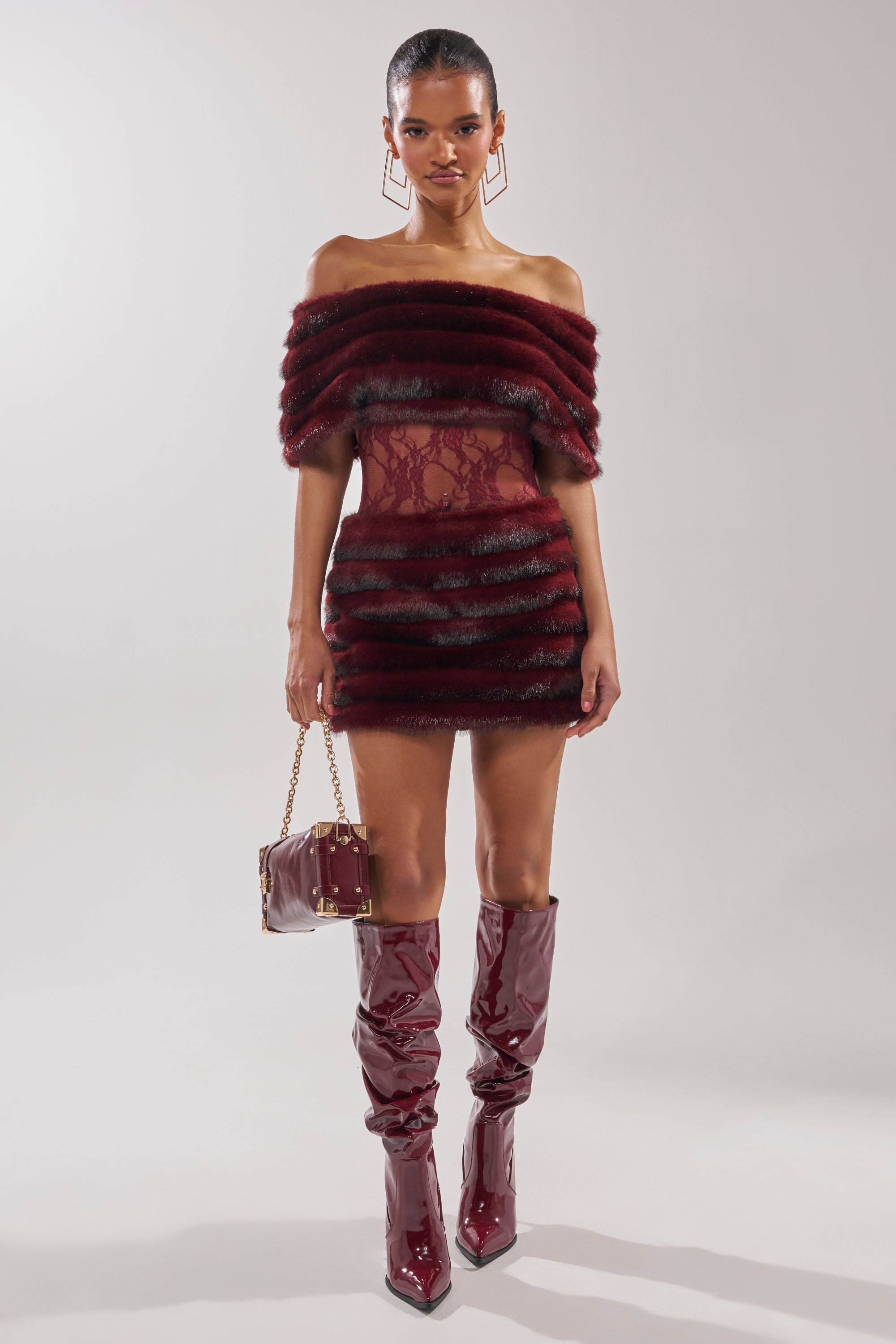 A woman poses against a plain background in the SOFT CHERRY SLEEVELESS MINI DRESS, featuring burgundy faux-fur tiered panels and lace detailing, styled with matching knee-high boots and a coordinated handbag.