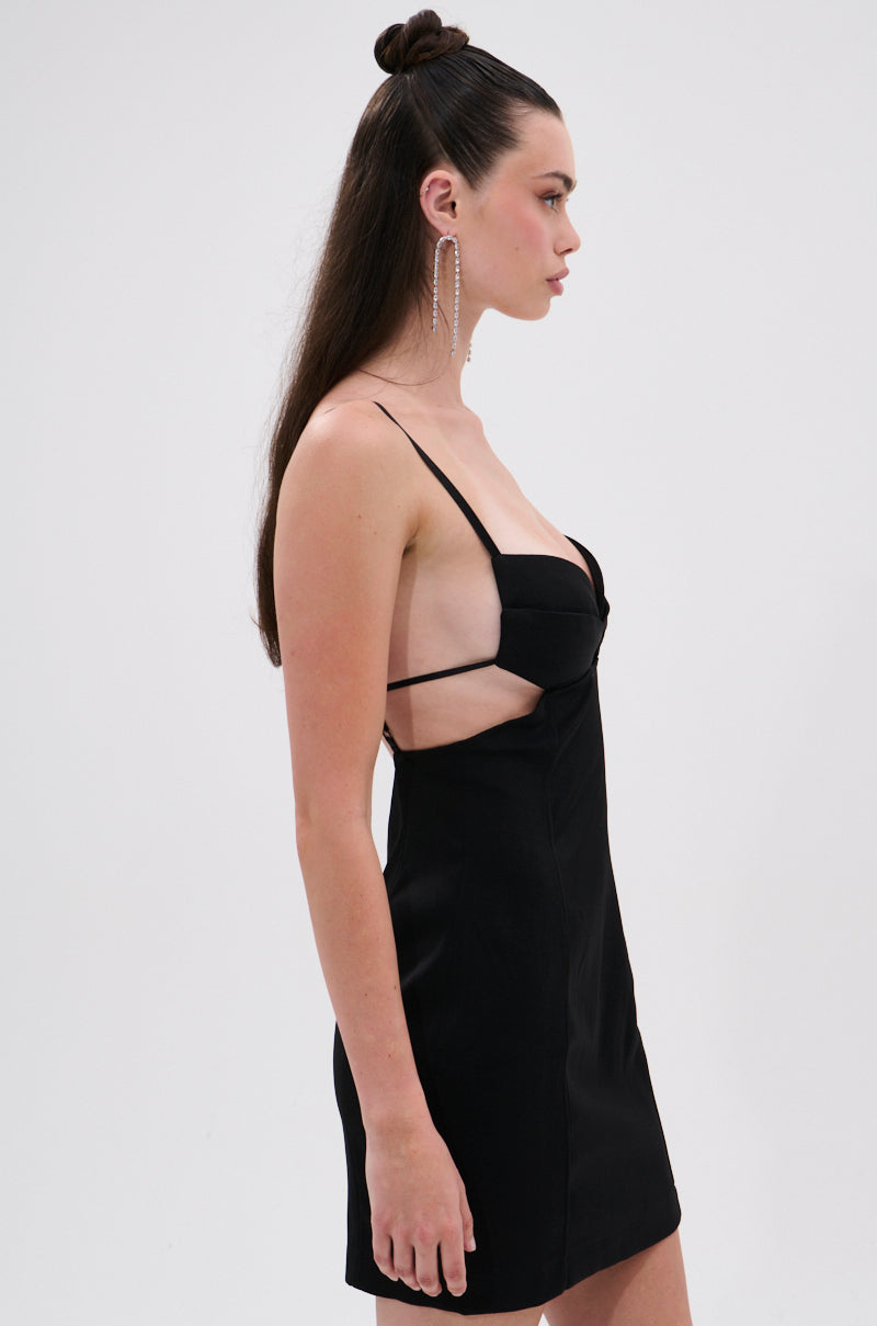 A woman with long dark hair in a half-up style wears the MIAMI NIGHTS MINI DRESS WITH CUTOUTS—a sleeveless black mini with thin straps, side cutouts, and a curve-hugging fit—standing in profile against a plain white background.
