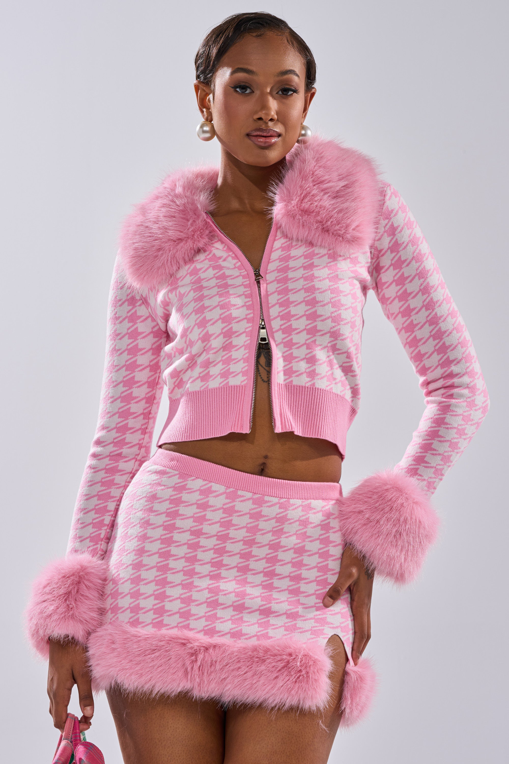 A woman models the QUEEN OF LOVE SWEATER WITH FUR TRIM, featuring bubblegum pink and white houndstooth with faux fur accents at the collar, cuffs, and hem. She accessorizes with hoop earrings and a coordinating pink bag.