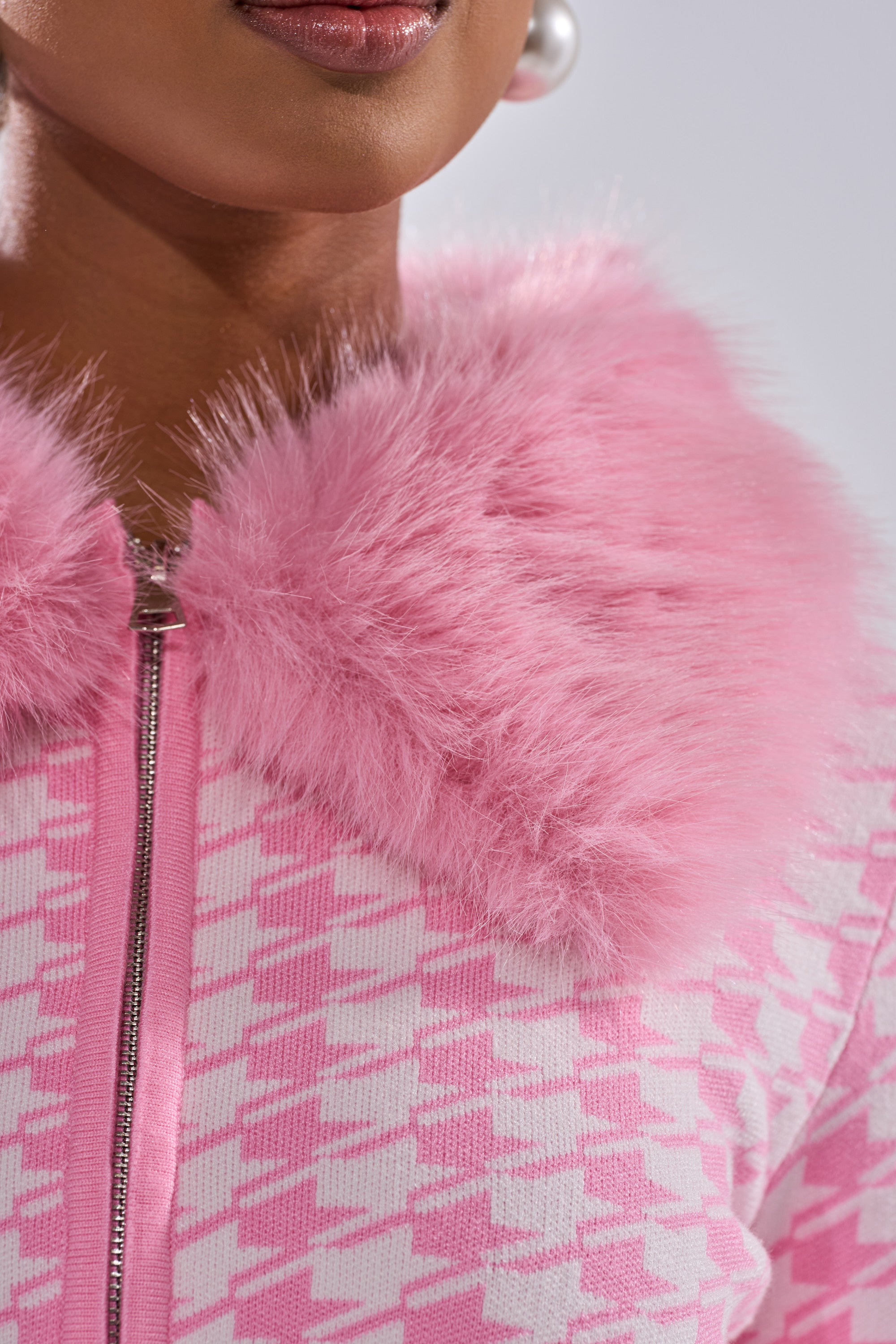 A close-up shows a person in the QUEEN OF LOVE SWEATER WITH FUR TRIM, featuring a pink and white houndstooth pattern, fluffy faux fur collar, silver zipper, and a partially visible large pearl earring.
