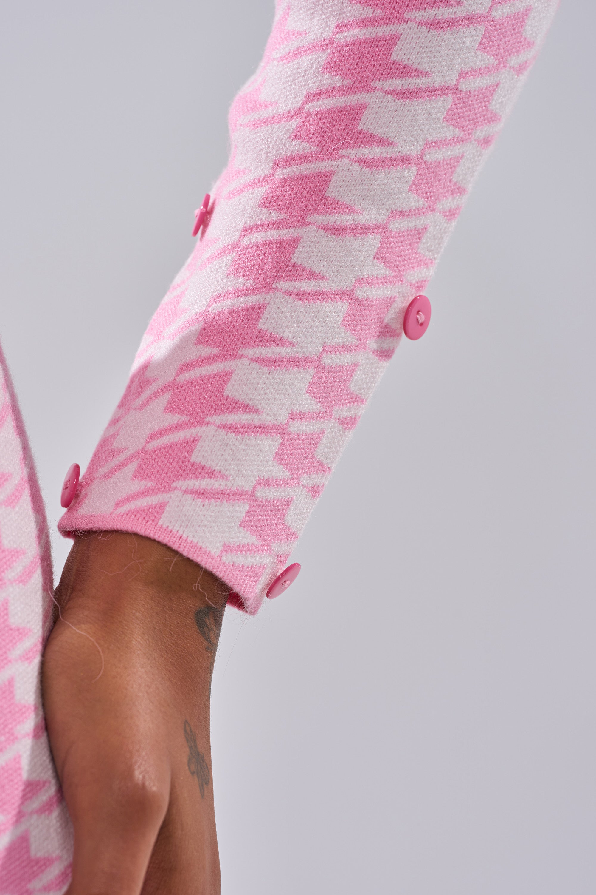 A close-up of a person's arm in the QUEEN OF LOVE SWEATER WITH FUR TRIM, featuring a bubblegum pink houndstooth knit sleeve with pink buttons. The hand is partially visible with a small wrist tattoo.