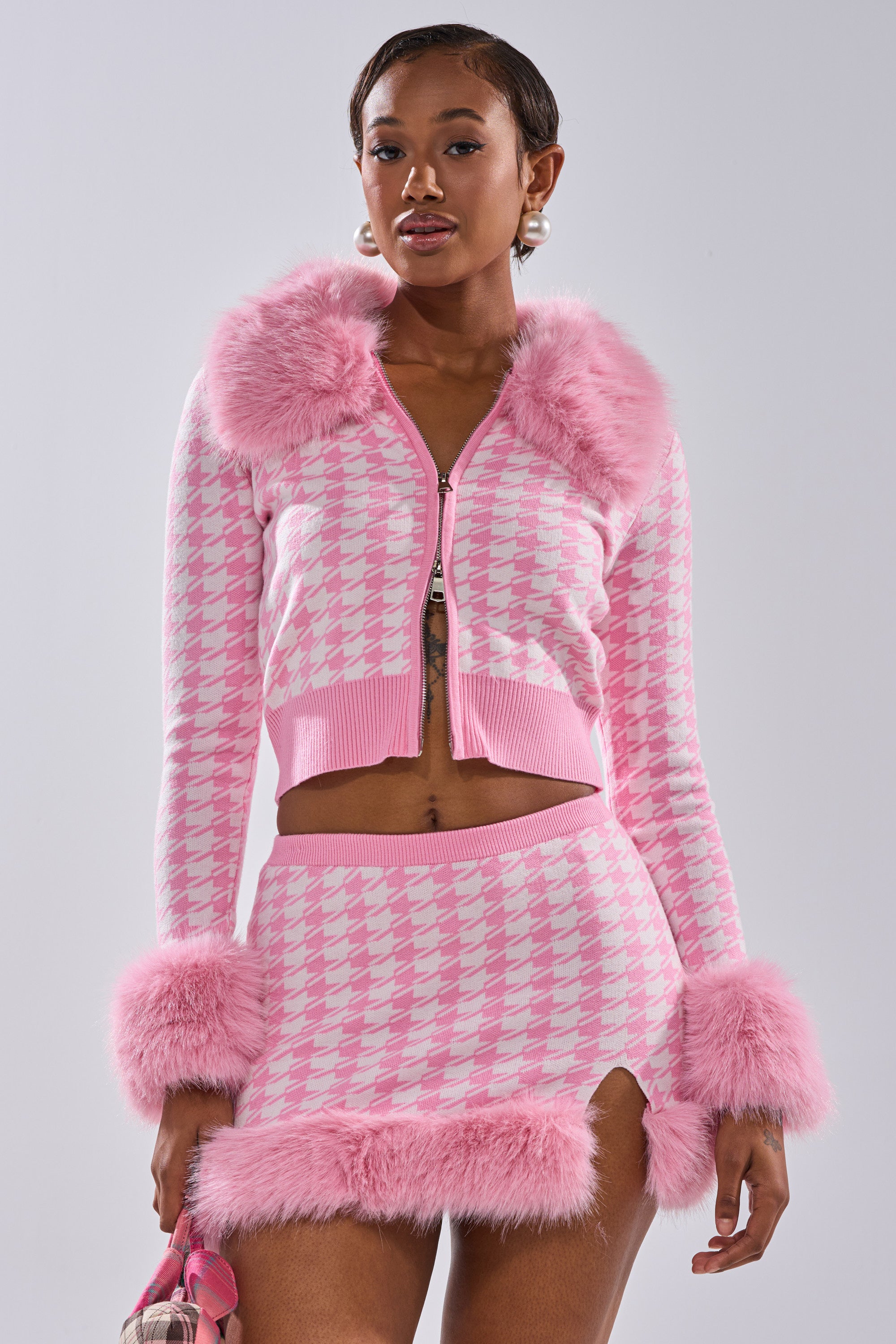 A woman wears the QUEEN OF LOVE SWEATER WITH FUR TRIM, a bubblegum pink cardigan with fluffy faux fur at the collar, cuffs, and hem, paired with a matching houndstooth mini skirt for a playful, coordinated look.