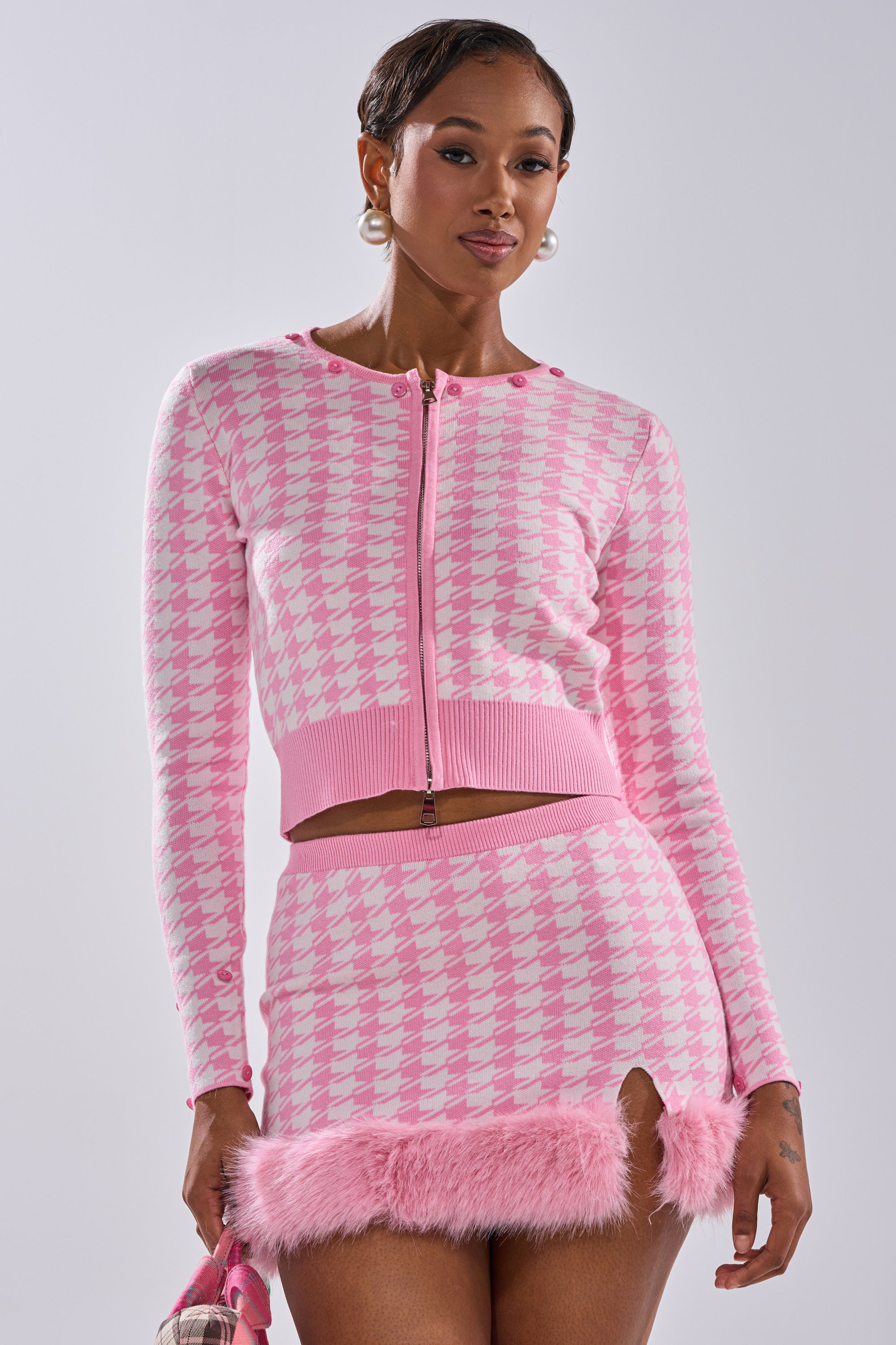 A woman models the QUEEN OF LOVE SWEATER WITH FUR TRIM in pink and white, paired with a matching mini skirt and handbag. She accessorizes with large hoop earrings, standing against a plain light background for a chic look.